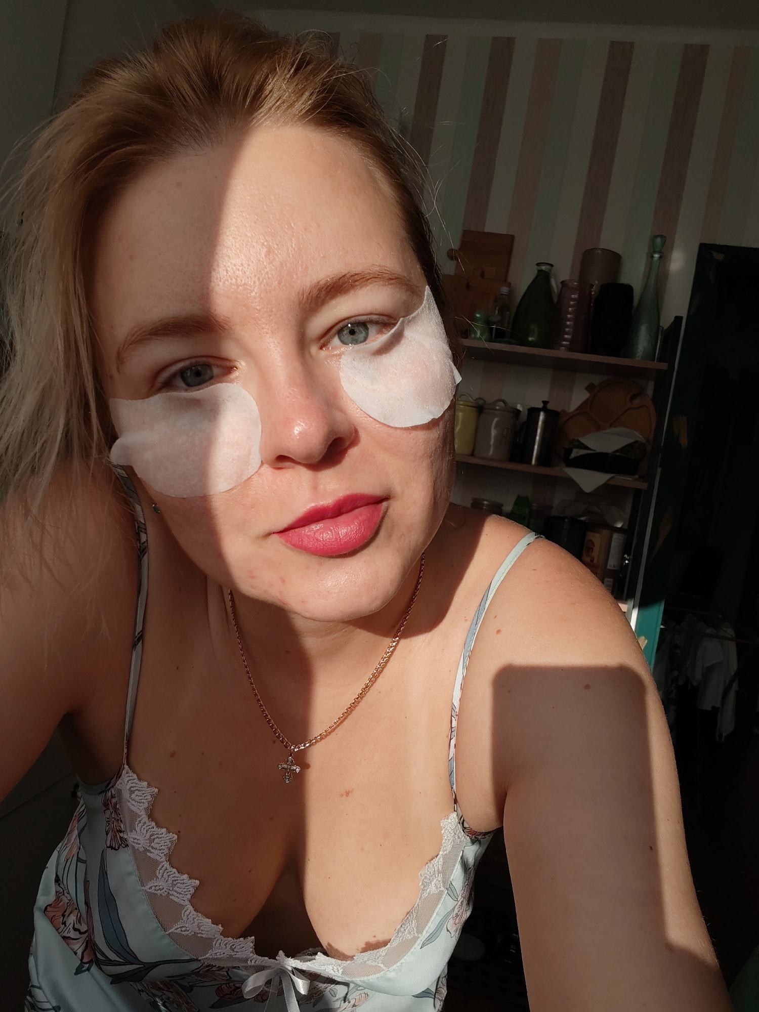 The image shows a close-up of a woman with blonde hair and blue eyes. She is wearing a light blue camisole with white lace trim and a gold necklace with a cross pendant. White under-eye patches are applied to her skin. The background consists of a wall with vertical striped wallpaper and shelves filled with various items, including bottles and jars. Sunlight casts a shadow across the woman's face. The setting is likely a domestic environment. There is no indication of the specific location within Izhevsk, Russia.