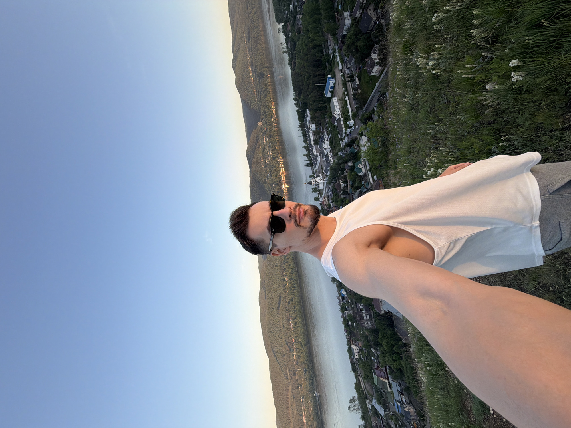 A man is captured in a selfie-style photograph, positioned on a grassy hillside overlooking a panoramic landscape in Krasnoyarsk, Russia. He is depicted from the mid-chest up, facing slightly to his right, with a dark beard, short dark hair, and wearing black sunglasses. He is dressed in a white sleeveless tank top and partially visible grey sweatpants. His right arm, holding the camera, extends into the lower left foreground. The background features a wide river, likely the Yenisei, flowing horizontally across the mid-ground. Along the riverbanks, a settlement of houses and buildings is visible, including a prominent white complex with domes, consistent with a monastery. Dense forested hills rise beyond the river. The immediate foreground shows green grass with scattered white blossoms. The sky is clear and light blue, illuminated by warm, golden-hour sunlight that highlights the man's left side.