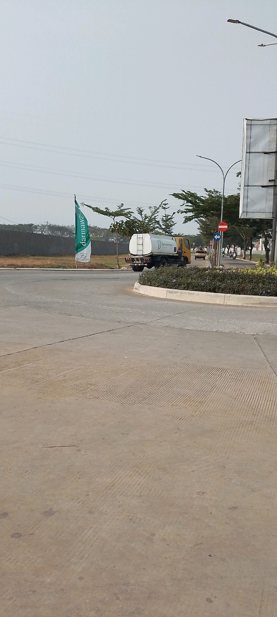 This is a medium-shot, eye-level, outdoor photograph taken on a bright, overcast day in Satriamekar, Indonesia. The image captures a road intersection within what appears to be a new development or industrial area, with a yellow water truck positioned on the road, turning. The truck has a white tank with the word "darmawatesa" visible. To the left of the truck, a tall, green and white flag with "darmawa" is planted in the ground. In the background, there is a concrete wall, some trees, and a few houses. A white sign is visible on the right side of the frame. The road in the foreground is paved concrete with visible expansion joints. There are streetlights visible in the upper right portion of the image. A few motorcycles and other vehicles are seen in the distance on the road behind the water truck. The overall environment suggests a relatively new or under-construction area.