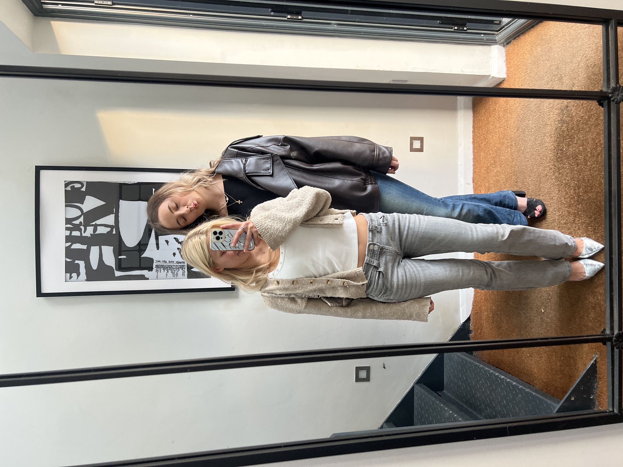 Two women are lying on the floor, posing for a mirror selfie in an indoor environment. One woman, closest to the mirror, has blonde hair and is holding a smartphone while wearing a white tank top, a beige cardigan, and grey jeans. The other woman has long brown hair and is wearing a black shirt, a brown leather jacket, and blue jeans. A framed black and white poster is on the wall behind them. The floor has two types of coverings, one is a beige carpet and the other is a gray metal stairwell. The reflection is framed by a black metal-framed mirror. The location appears to be an urban setting, possibly London, United Kingdom, given the architectural style.