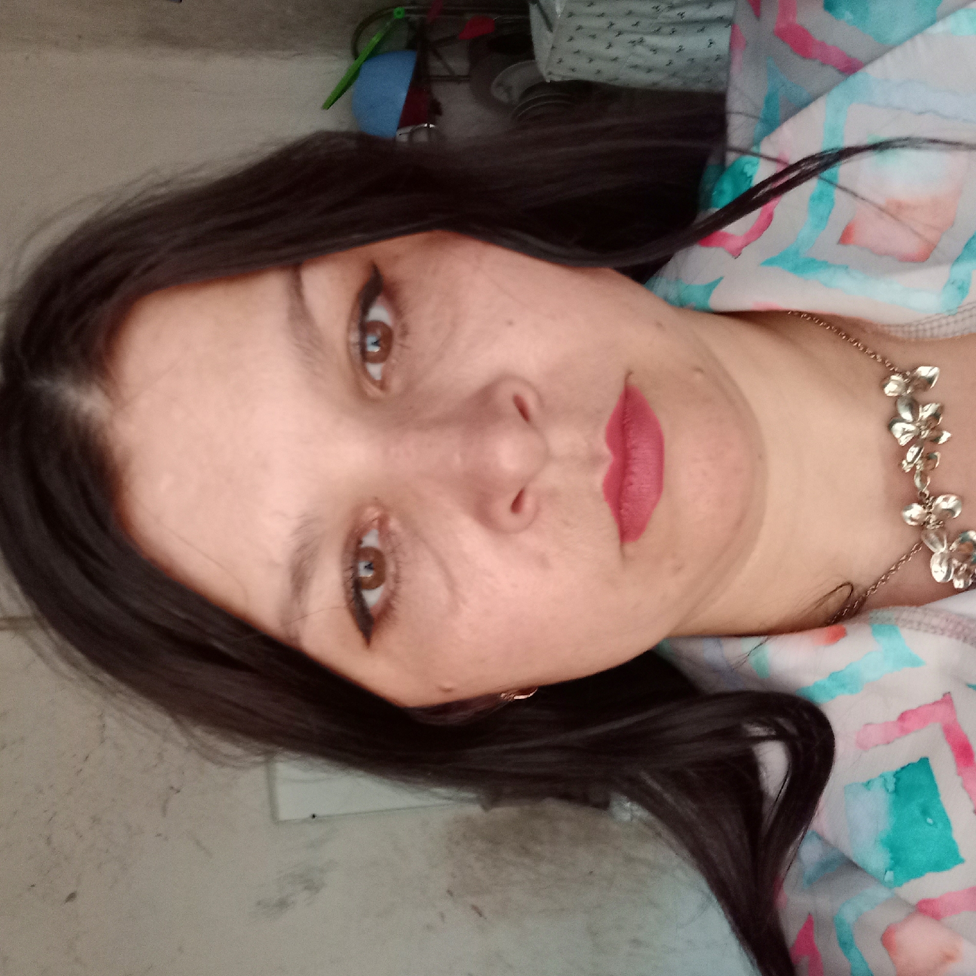 The image is a selfie of a woman with dark hair, brown eyes, and pink lipstick. She is wearing a patterned shirt and a flower-shaped necklace. The background appears to be an indoor setting with a textured wall. The location is identified as Klyntsi, Ukraine.