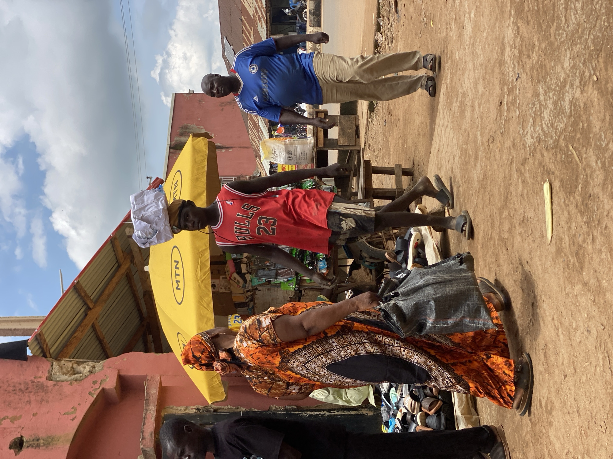 In Kafanchan B, Nigeria, a busy street scene unfolds under a partly cloudy sky. In the foreground, a woman in a vibrant orange and black patterned dress, her head covered with an orange cloth, carries a large gray bag. She walks alongside a young man wearing a red basketball jersey with the number 23 and distressed shorts, a white package balanced on his head. To their right, another man in a blue t-shirt and khaki pants walks away from the viewer. Behind them, a yellow market umbrella with the "MTN" logo provides shade over a stall displaying various items, including shoes and bags. The ground is a dusty, reddish-brown dirt. In the background, buildings with corrugated metal roofs and faded paint line the street, suggesting a bustling marketplace or commercial area.