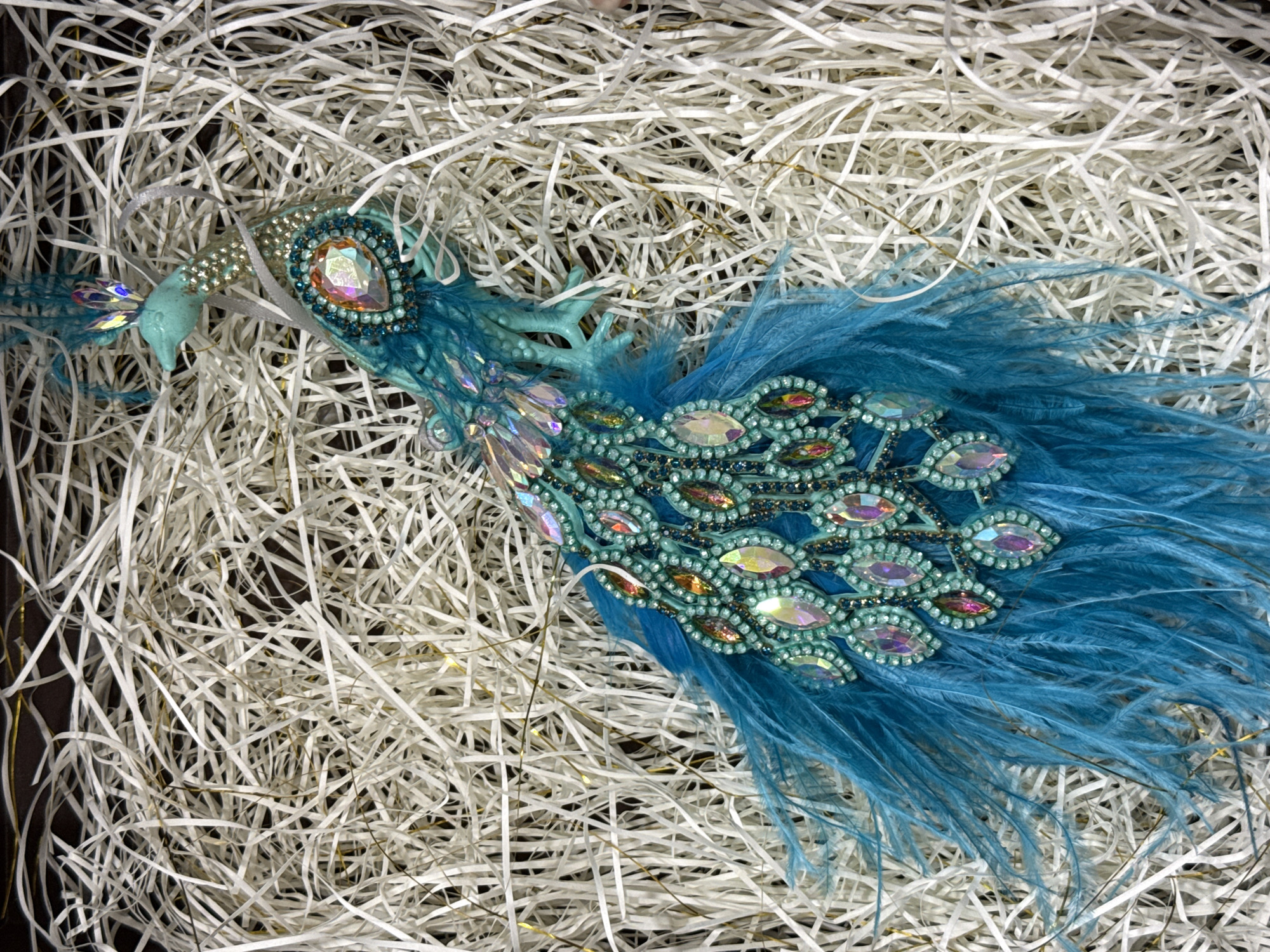 The image shows a decorative peacock ornament lying on a bed of white shredded paper. The peacock is predominantly teal and blue, adorned with numerous iridescent rhinestones and jewels. It has a long, fanned tail made of blue feathers and intricate detailing along its body and neck. The ornament appears to be an artificial creation, possibly a Christmas tree ornament or a craft item. The background consists of a dark surface partially obscured by the white shredded paper. There is no discernible location in the city of Dnipro, Ukraine, or any indication of human presence.