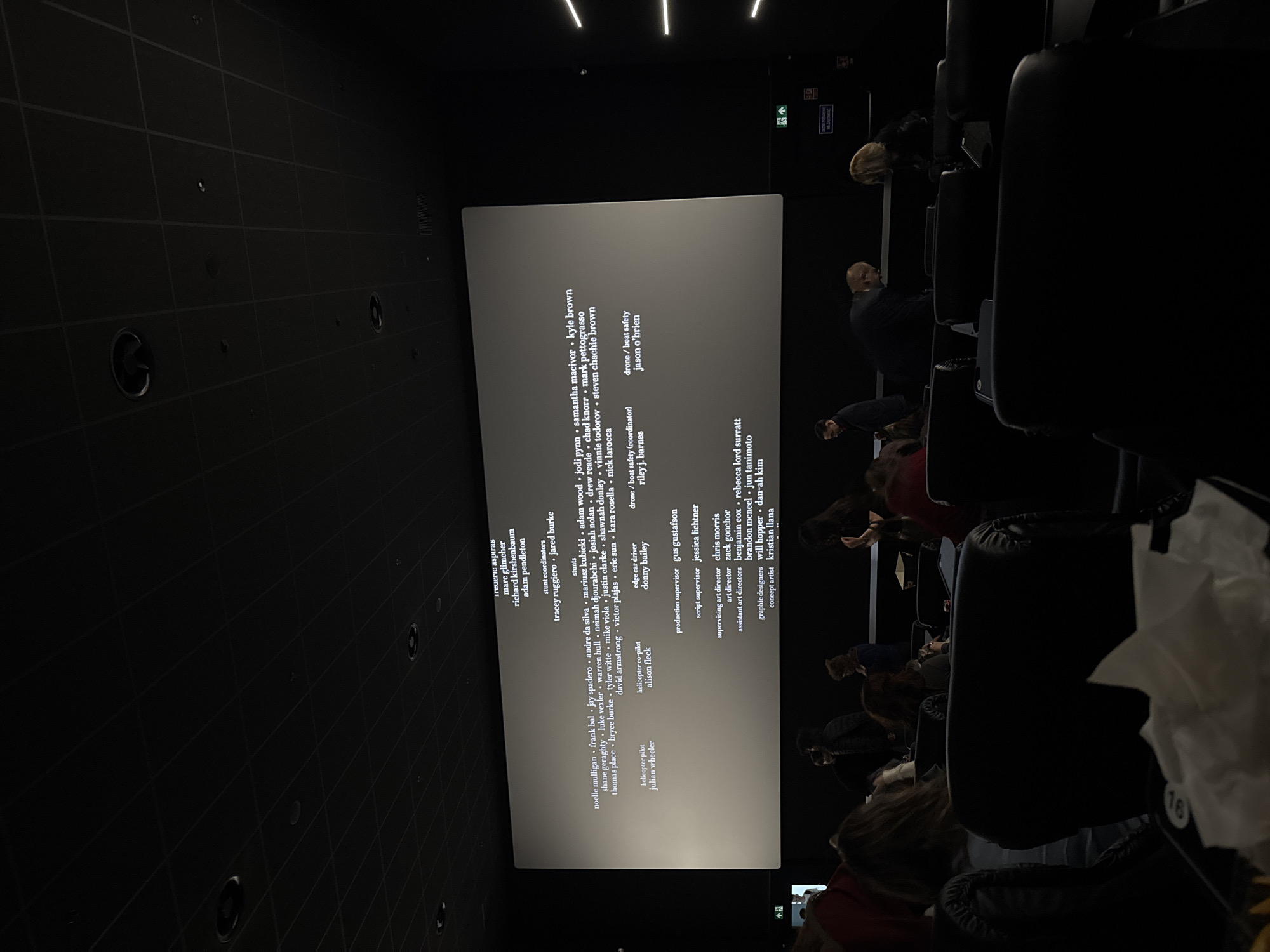 The image shows a darkened movie theater with people sitting in rows of seats, facing a large screen displaying the end credits of a film. The names of the people involved in the production of the film are on the screen. The environment is dark, typical of a movie theater during a showing.