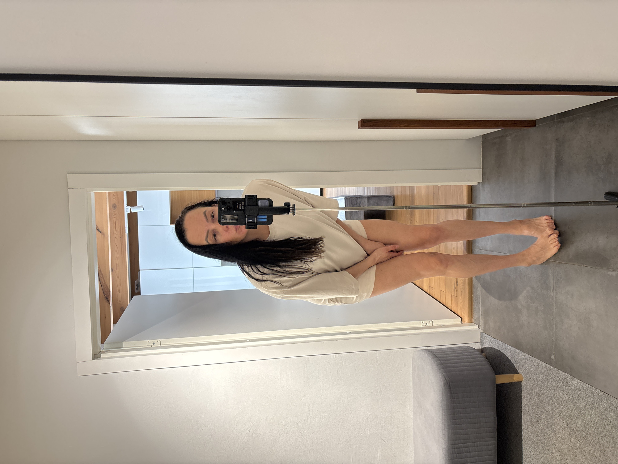 A woman is taking a selfie in a mirror. She is using a selfie stick attached to her phone. She is wearing a beige top. Her hair is long and dark. She is barefoot. The background shows a room with a bed, wooden floors, and a window. The image was likely taken in Chaikovsky, Russia.