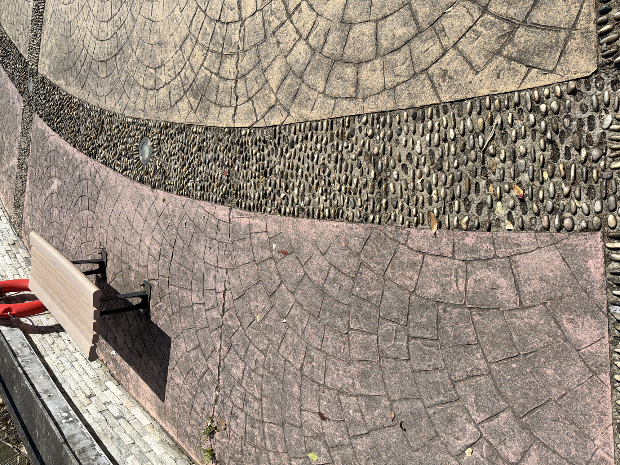 The image shows a section of a walkway or patio area. The ground is paved with three distinct sections: a beige-colored section on top, a strip of small pebbles in the middle, and a pink-tinted section on the bottom. The paving pattern on the beige and pink sections consists of semi-circular blocks arranged in an arc. There are several dead leaves scattered across the walkway.
A bench, attached to the pink-paved area, is visible in the lower left. The bench has a wooden seat and black metal supports. A red plastic object is partially visible next to the bench.
Two small circular lights are embedded in the pebble strip. The location is presumed to be Jiangning District, China. There are no people visible in the image.
