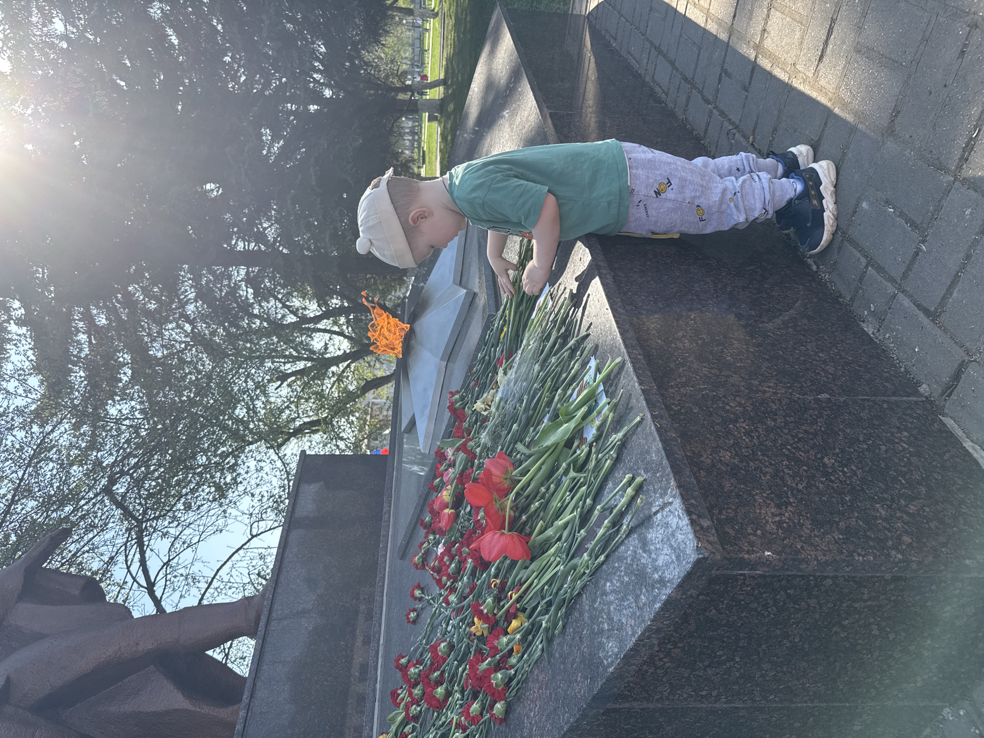 A young child lays red carnations on a dark granite memorial in Zav'yalovo, Russia. The child, wearing a green shirt, gray pants, a light-colored hat with pom-poms, and dark blue sneakers, leans over the top of the memorial, apparently arranging the flowers. A flame burns inside the star-shaped element of the memorial. Several bouquets of red and yellow flowers are spread on the monument's top surface. The monument sits on a paved area bordered by greenery and trees. Part of a dark brown sculpture of human arms is visible. Bright sunlight glares in the upper left of the image.