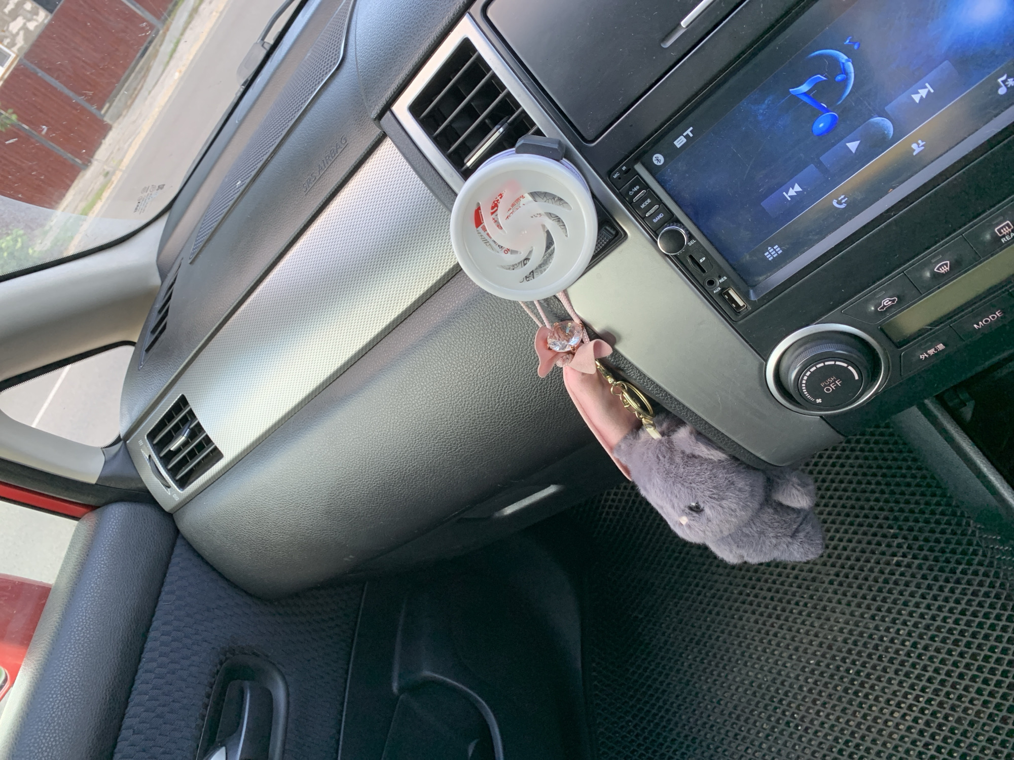 The image shows the interior of a car. In the foreground, a grey fuzzy keychain resembling an elephant is hanging from the dashboard. Next to it is a white car air freshener with a swirl design. The dashboard has a silver accent strip and an air vent. Below the air freshener is a car stereo with a touchscreen display showing music controls. To the left of the stereo is a dial with "PUSH OFF" labeled on it. The car's floor mat is visible in the lower left corner. The scene appears to be in the city of Belogorsk, Russia, as indicated by the context.