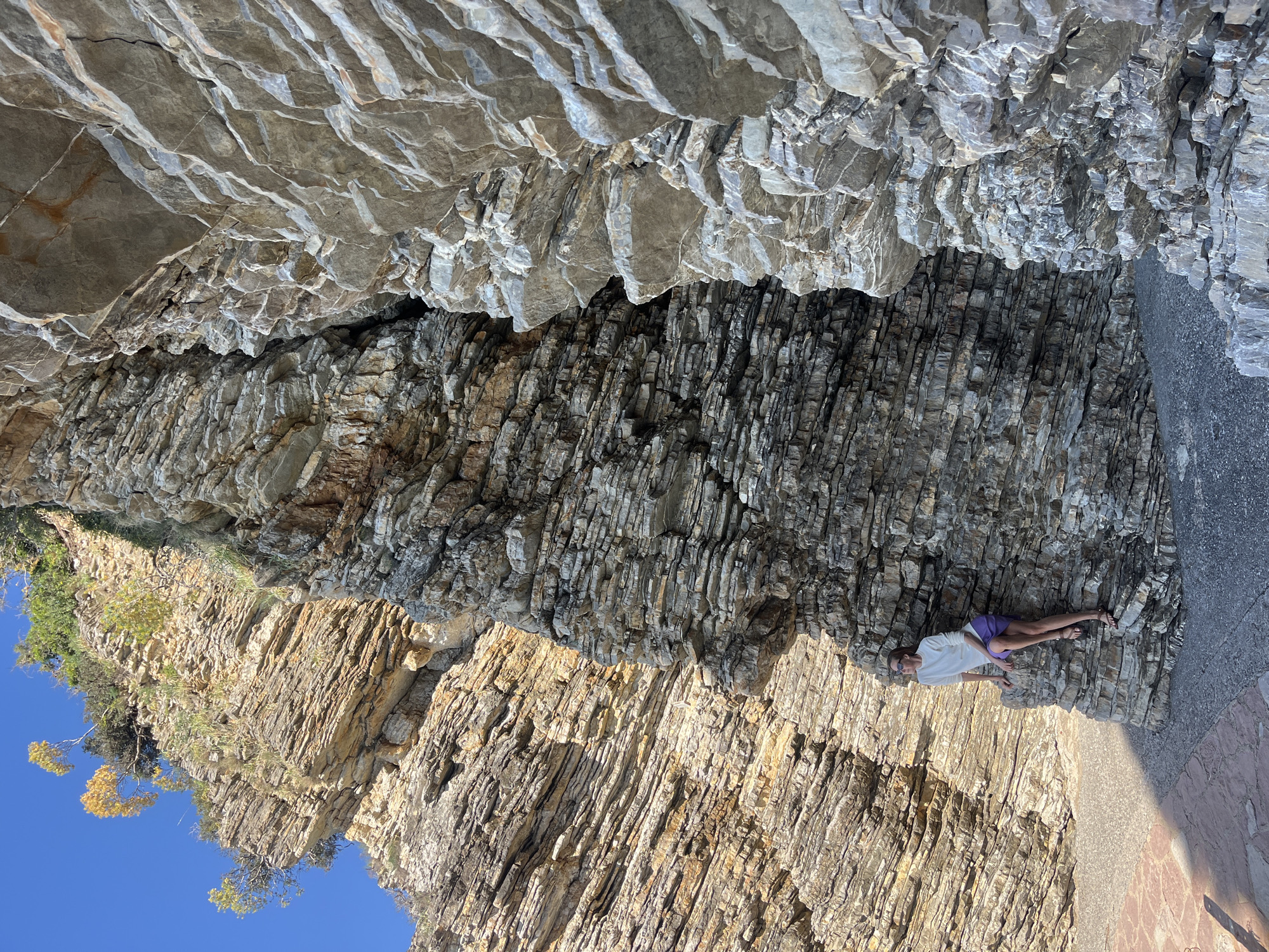 A vertically oriented image shows a person lying on a rock formation near Budva, Montenegro. The rock face consists of stratified layers of light brown and grey rock. The person, wearing a white shirt and purple shorts, is positioned horizontally along a ledge of the rock. The lower right corner of the image shows a paved surface. The sky is visible in the lower left, indicating a sunny day.