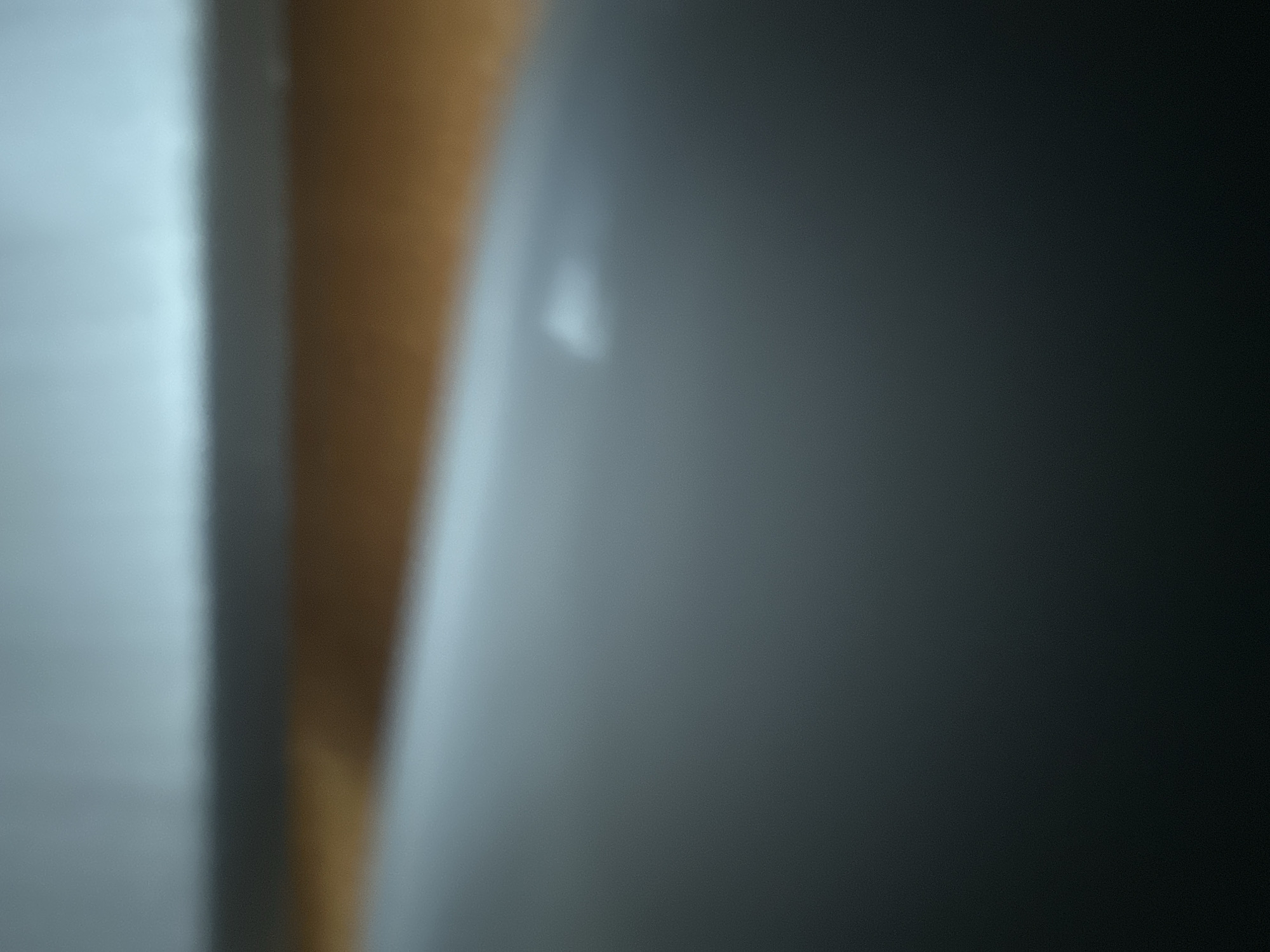 This is an abstract, out-of-focus image with a shallow depth of field. The dominant elements are broad strokes of light and shadow. On the left side of the image, a bright, diffused light illuminates a pale blue or white area, with a vertical band of a warm, light brown color running through the center. This brown band is darker towards the right. To the right of the brown band, the image transitions into a gradient of dark gray and black, with a small, blurry, brighter spot near the center-left of this dark area. The overall impression is of light interacting with indistinct shapes, possibly indicating surfaces or objects in a dimly lit environment. There are no discernible people, specific objects, or clear setting details. The abstract nature makes it impossible to identify any activity, time of day, weather, location cues, or emotions. There is no visible text in the image.
