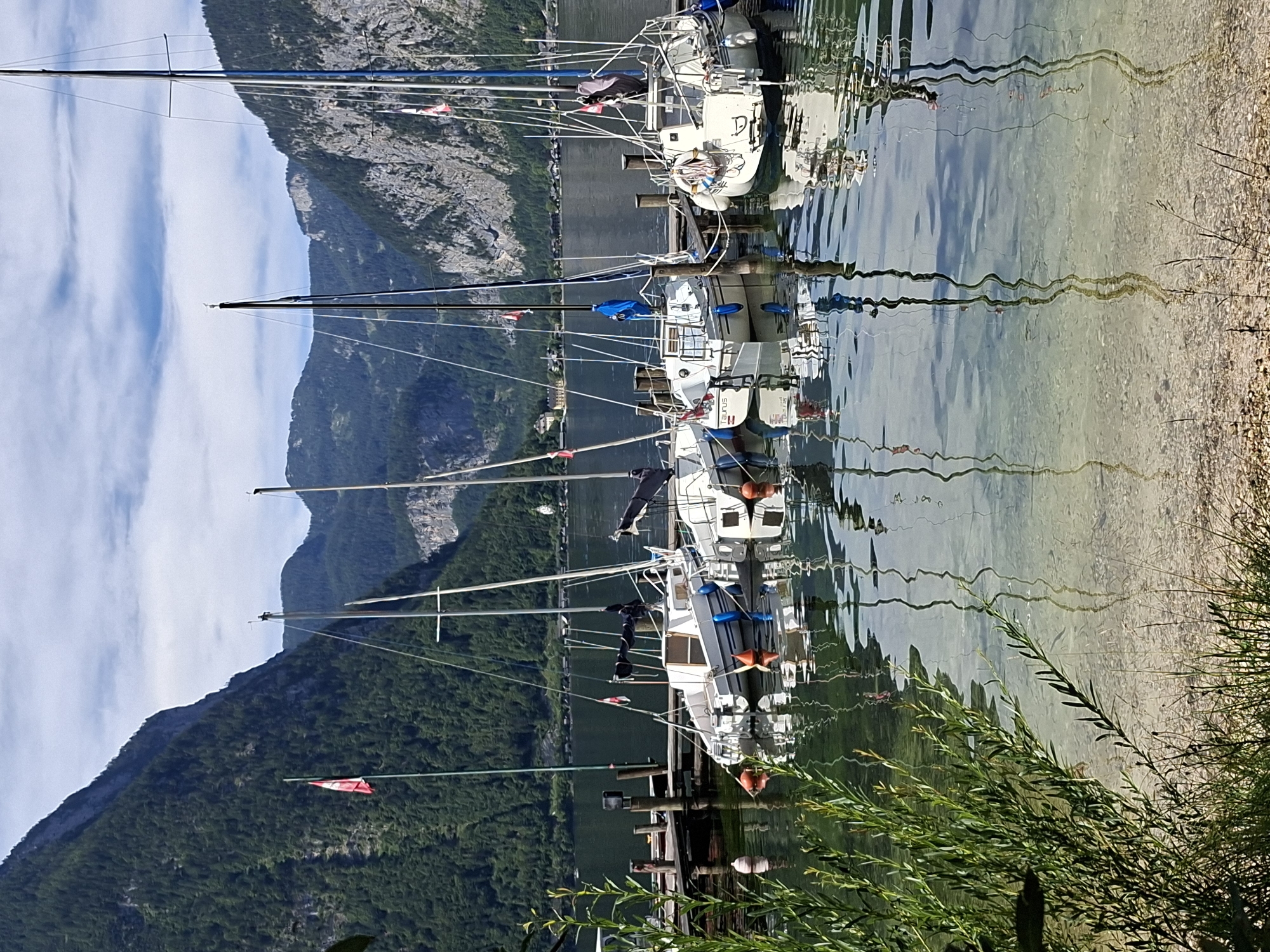 This image depicts a serene harbor in Obertraun, Austria, nestled at the foot of majestic, tree-covered mountains. The foreground is dominated by the tranquil waters of a lake, reflecting the sky and the surrounding scenery. Along the shore, a wooden dock is lined with several sailboats, their masts reaching towards the sky. The boats, mostly white with some blue accents, are moored and appear to be at rest. Orange buoys are visible in the water, marking the dock and possibly indicating mooring spots. A small, red and white flag is attached to a flagpole on the dock. The background is defined by the imposing mountains, with a clear blue sky dotted with white clouds. The overall atmosphere is one of peacefulness and natural beauty.