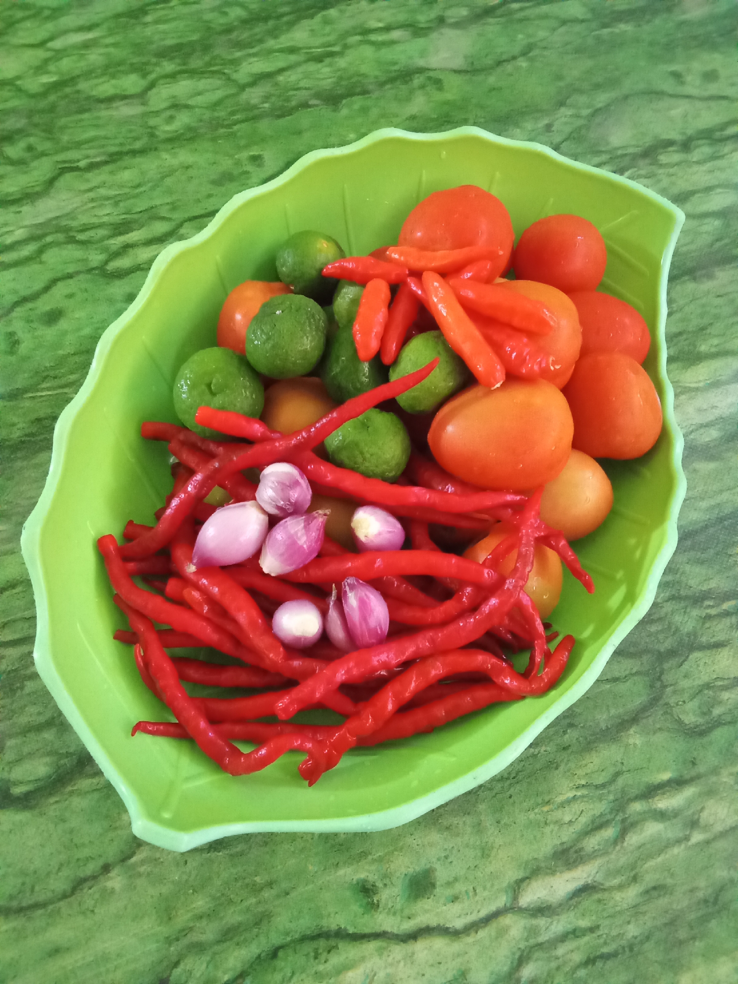 A bright green, leaf-shaped plastic bowl contains a colorful assortment of fresh ingredients on a green, marble-patterned surface. The bowl is filled with several red chilies, some long and thin, others shorter and fatter. There are also several small, round tomatoes, their skins a vibrant orange-red. Interspersed among the tomatoes and chilies are small, green citrus fruits, likely limes. A few small, peeled shallots, their purplish-white layers visible, are also present. The ingredients are arranged loosely within the bowl, suggesting they are prepared for cooking. The background is a textured green surface that resembles polished marble or stone, with darker green veins running through it. The lighting is even, casting no strong shadows, indicating an overhead light source. The overall impression is one of fresh produce ready for culinary use, set against a clean, visually appealing backdrop. The image does not depict any specific location in Subang, Indonesia, beyond what can be inferred from general food preparation settings common in many regions.