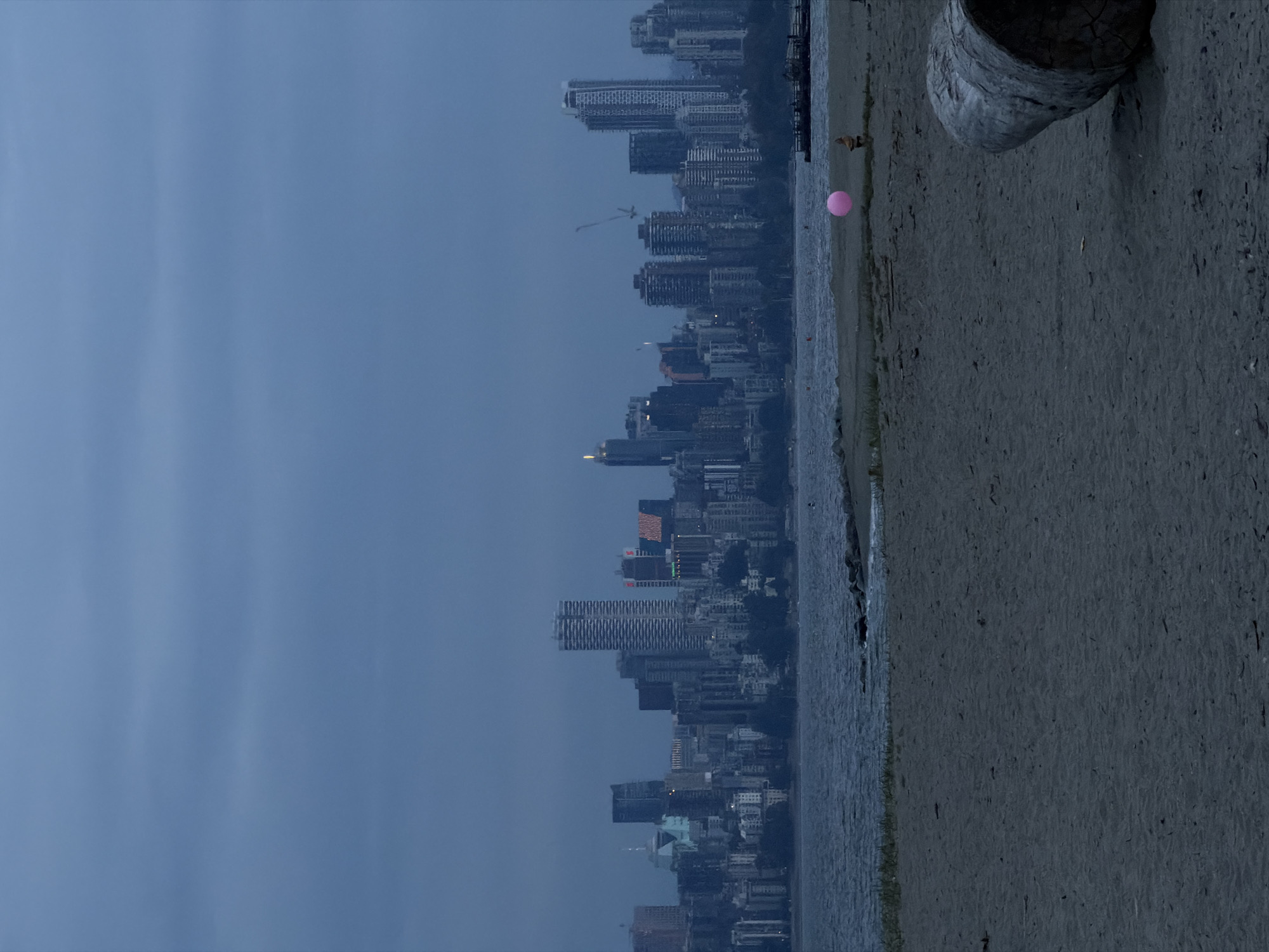 This is a wide-angle, eye-level, low-light, over-the-shoulder shot of a beach in Vancouver, Canada, with the city skyline in the background. The sky is overcast and a deep blue, with some horizontal cloud striations. The water is choppy, a dark blue, and meets a sandy beach in the foreground. A large piece of driftwood is in the lower right quadrant of the image. In the middle ground of the beach, a person is standing and a bright pink balloon is floating in the air, to the left of the person. The city skyline, dominated by numerous skyscrapers of varying heights and architectural styles, stretches across the horizon. The overall mood of the image is somber and tranquil, likely captured at dusk or dawn due to the low light conditions.