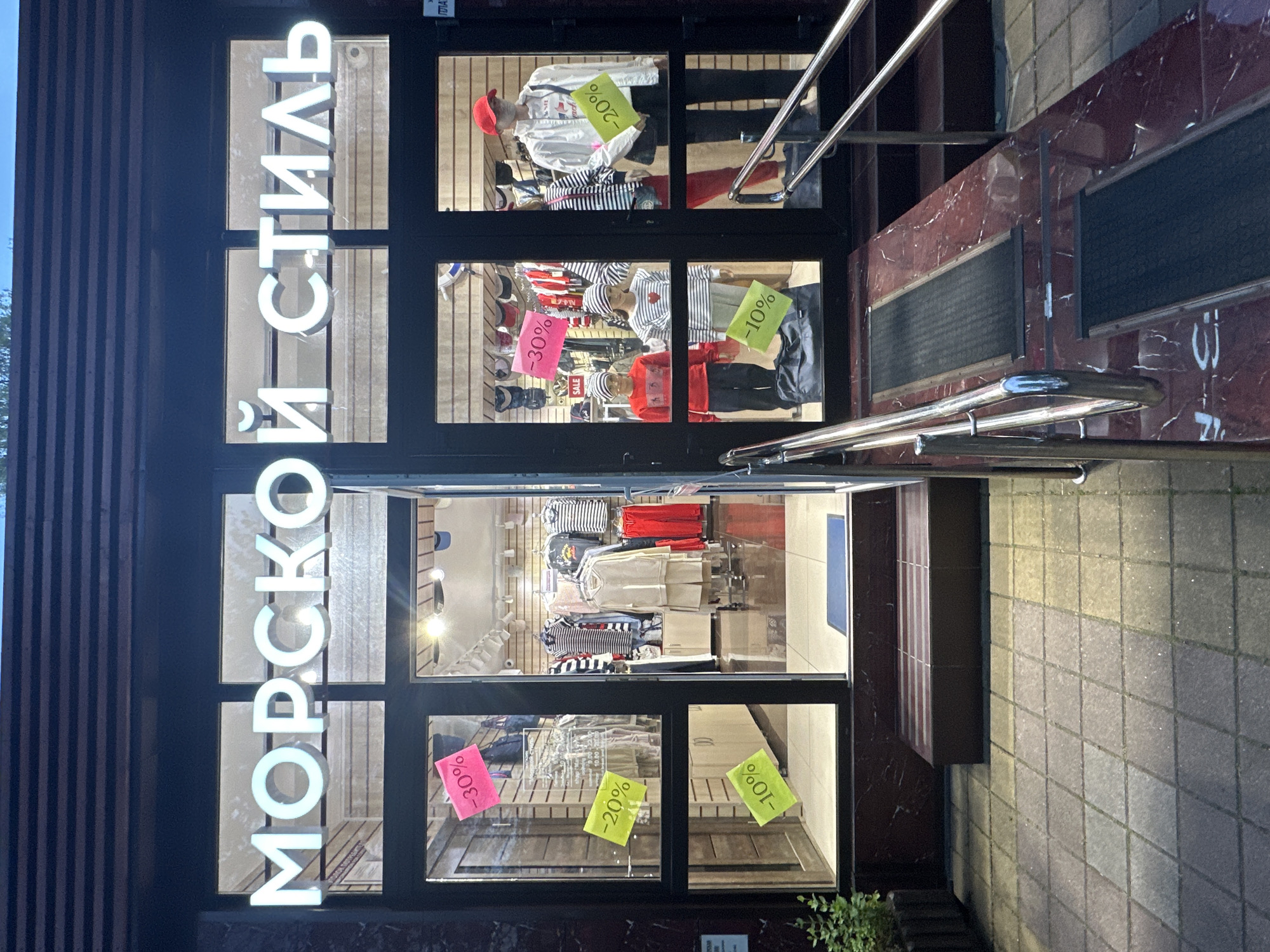 The image displays the exterior of a clothing shop named "МОРСКОЙ СТИЛЬ" (MORSKOY STIL), which translates to "Marine Style" or "Sea Style," in Sochi, Russia. The shop's name is rendered in large, bright white, illuminated Cyrillic letters positioned vertically along the left side of the entrance.
The storefront consists of large display windows, through which various clothing items are visible. Mannequins model outfits, including striped shirts, red tops, a white jacket with a red hat, and other apparel, indicative of a casual style. Numerous neon-colored discount signs are affixed to the windows, advertising sales with prominent percentages: "-30%" (on pink signs), "-20%" (on yellow-green signs), and "-10%" (also on yellow-green signs). A small sign also reads "SALE". The interior of the shop appears brightly lit, showcasing racks of clothes and accessories.
A set of steps, made of dark reddish-brown material with a marble-like pattern, leads up to the shop's entrance. These steps are equipped with silver-toned metal handrails and black non-slip strips. The ground level surrounding the shop consists of square paving stones. The scene appears to be set during the day, with natural light falling on the pavement, while the shop's interior lights and exterior sign remain illuminated. No people are visible in the image.