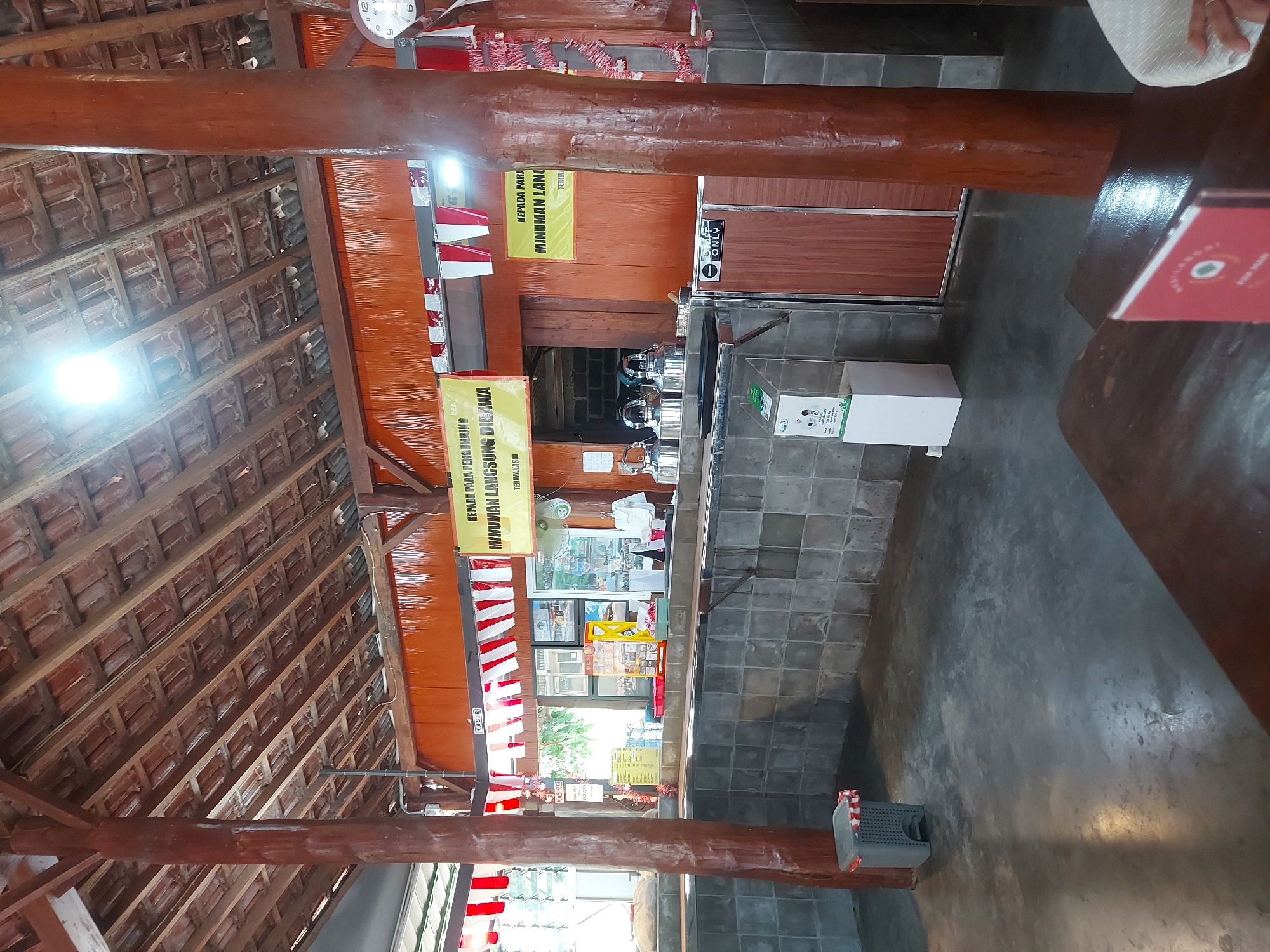 The image shows the interior of a food stall or restaurant with a rustic, open-air design, located in Kricak, Indonesia. The ceiling is made of exposed wooden beams and terracotta tiles. Red and white Indonesian flags are hung as decorations. In the background, a counter area is visible with several stainless steel kettles, a cash register, and some menus. There are signs in Indonesian that say "Kepada Para Pengunjung" (To the Visitors) and "Minuman Langsung Dibawa" (Drinks to be Taken Immediately). A water dispenser is also present. The floor is concrete. There are no visible people in the image.