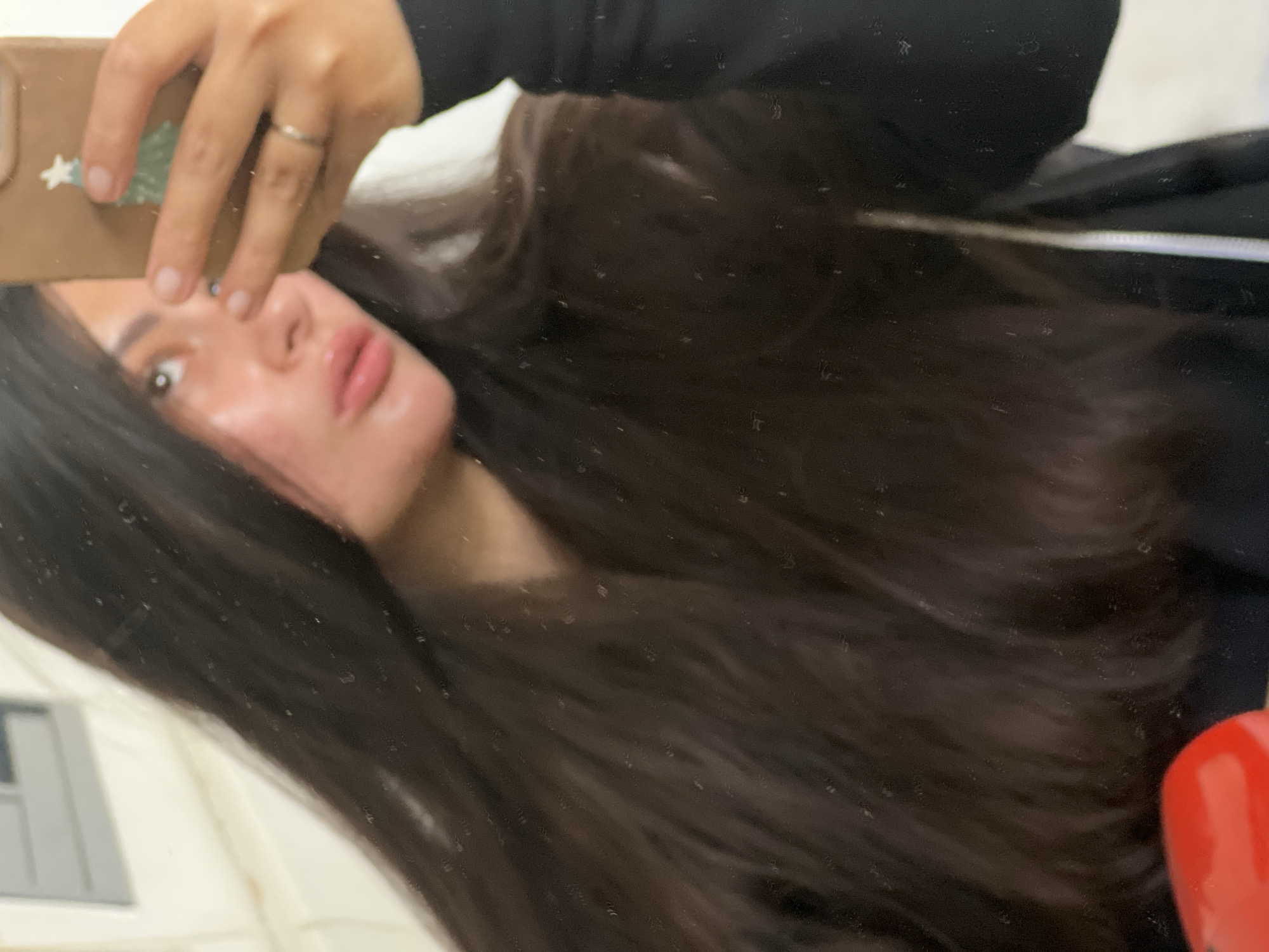 The image shows a person taking a selfie in a mirror. The person has long, dark brown hair and is wearing a dark-colored top. They are holding a phone with a light brown case. A silver ring is visible on their finger. The background includes a white wall and part of a red object. It's not possible to identify the location as Kiryat Bialik, Israel from the image.