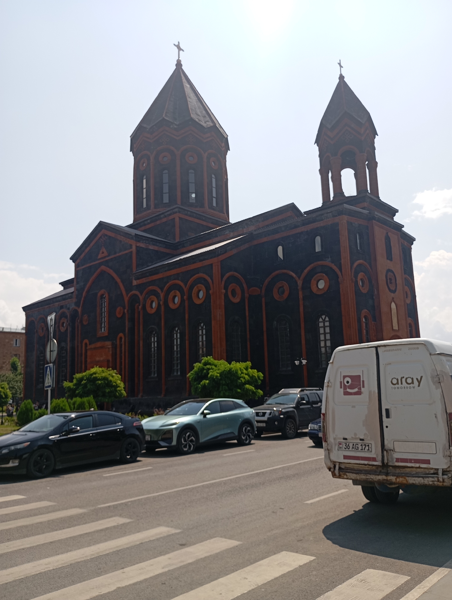 This is a medium shot, eye-level photograph taken on a sunny day in Gyumri, Armenia. The focal point of the image is the impressive Saint Mary's Cathedral, a prominent landmark constructed with dark volcanic rock and adorned with red tuff accents. The cathedral features two imposing towers, one of which is taller and capped with a cross. In front of the cathedral, a street is visible, featuring a pedestrian crossing in the foreground. Several cars are parked or driving on the street, including a black sedan, a light green SUV, and a white van with the "aray TOMORROW" logo. The background shows a clear, pale blue sky with a few scattered clouds. The overall impression is one of architectural grandeur and urban activity.