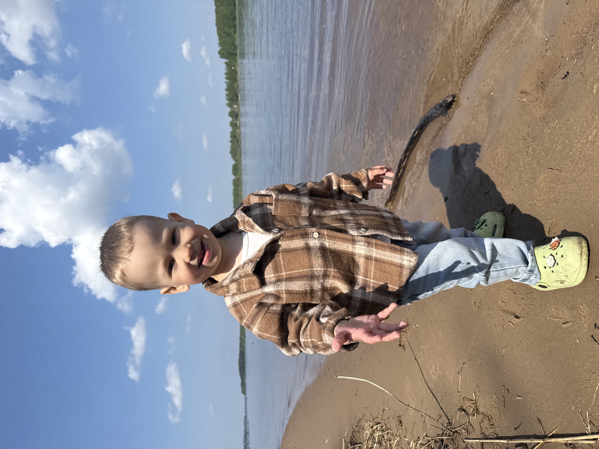 A young child stands on a sandy beach beside a body of water. The child is wearing a brown plaid shirt, a white t-shirt, blue jeans, and green clogs. The sky is blue with white clouds. A stick is lying on the beach near the water's edge. The location is Unknown, Unknown.