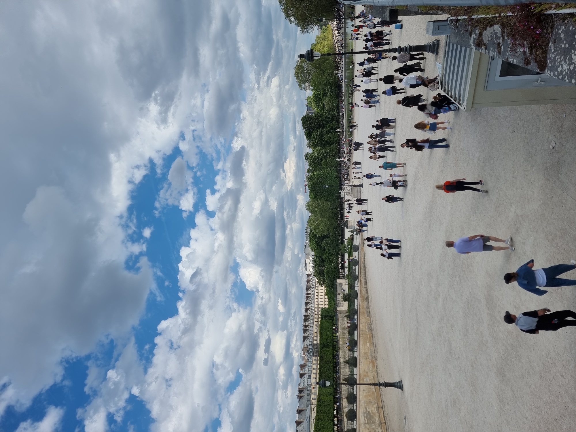 The image captures a scene in Paris, France. The perspective is from an elevated position, likely overlooking the Tuileries Garden. The scene shows a wide pedestrian walkway populated with numerous people, many strolling leisurely. A row of trees and hedges lines the walkway, separating it from the adjacent buildings and street. The buildings are visible in the background and appear to be typical Parisian architecture. The sky above is filled with clouds, with patches of blue sky visible in between. A light post is visible on the bottom left of the image. The people in the scene appear to be engaged in various activities, such as walking, talking, and relaxing.