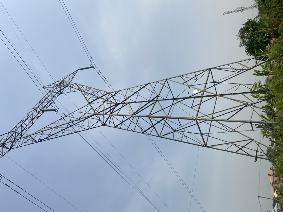 This image depicts a large, grey metallic lattice-work electricity transmission tower standing prominently against a clear, light blue sky. Multiple high-voltage power lines extend from the tower, suspended by green ceramic insulators visible in the upper section. The base of the tower is situated amidst dense green vegetation, including shrubs and what appears to be banana plants. In the mid-ground to the left, residential buildings with reddish-brown roofs and light-colored walls are partially visible, alongside a smaller utility pole. To the right in the distant background, another metallic lattice-work tower, likely a communication mast or another power pylon, is partially obscured by trees. No individuals are present. The scene is located in Fajo 2 Division Range, Nigeria, under bright daylight conditions.