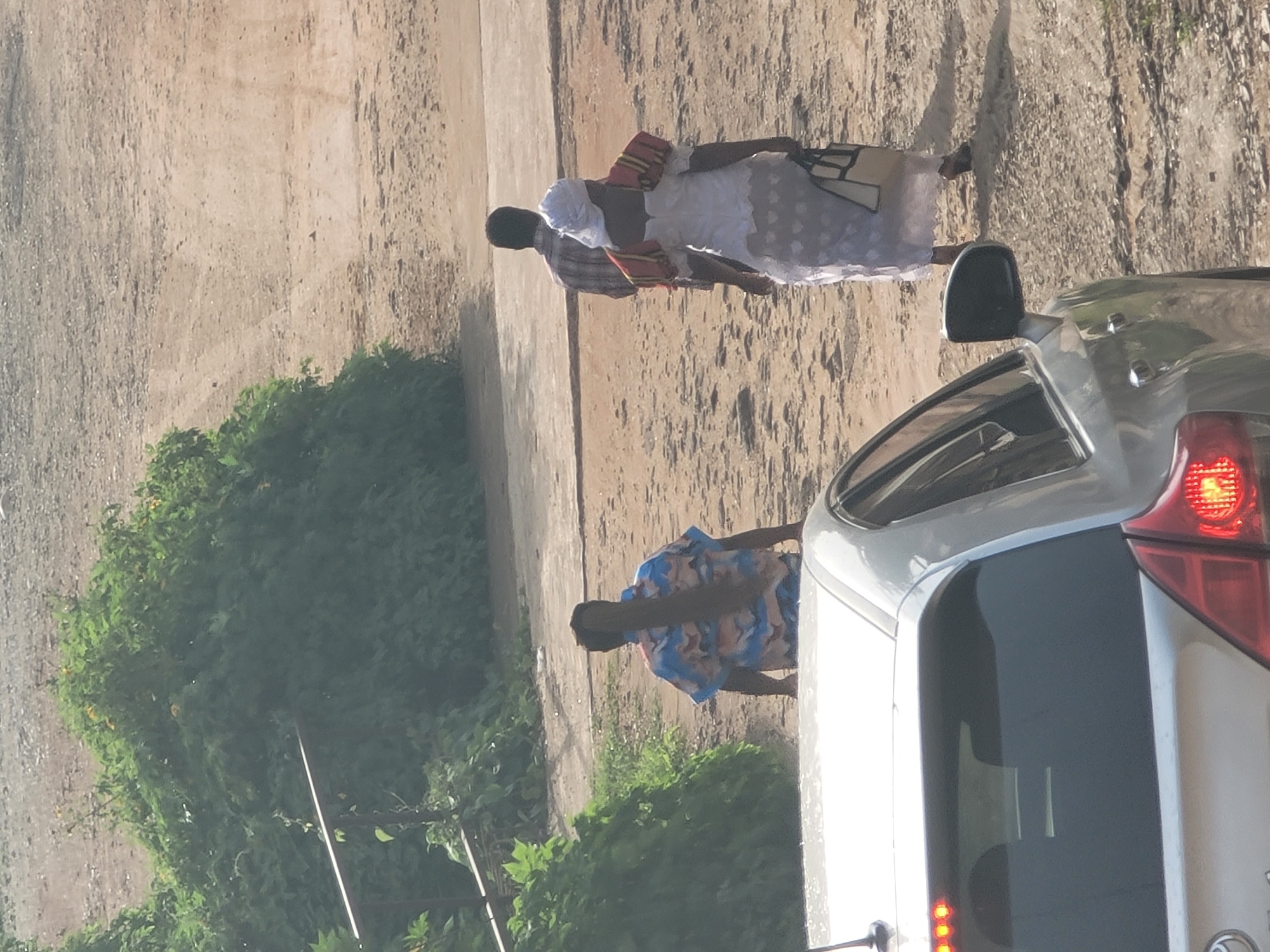 The image depicts a sunlit outdoor scene in Gura Top, Nigeria, focusing on a dirt path bordering a dense green bush. In the foreground, the rear of a silver car is visible, showing its tinted rear window and a portion of the left side of the vehicle. The car has red taillights illuminated. Further into the scene, three individuals are walking away from the camera. The first person, on the left, is a woman wearing a blue and pink patterned shirt and has very long, dark braided hair extending down her back. Next to her, slightly behind, is a person wearing a plaid shirt, their back to the camera. In the center right, a woman dressed in a white, ornate dress with red and yellow shoulder adornments and a white head covering is walking, carrying a light-colored bag with dark straps in her right hand. The ground is predominantly sandy dirt with some scattered debris and tire tracks, suggesting it's a frequently used pathway.