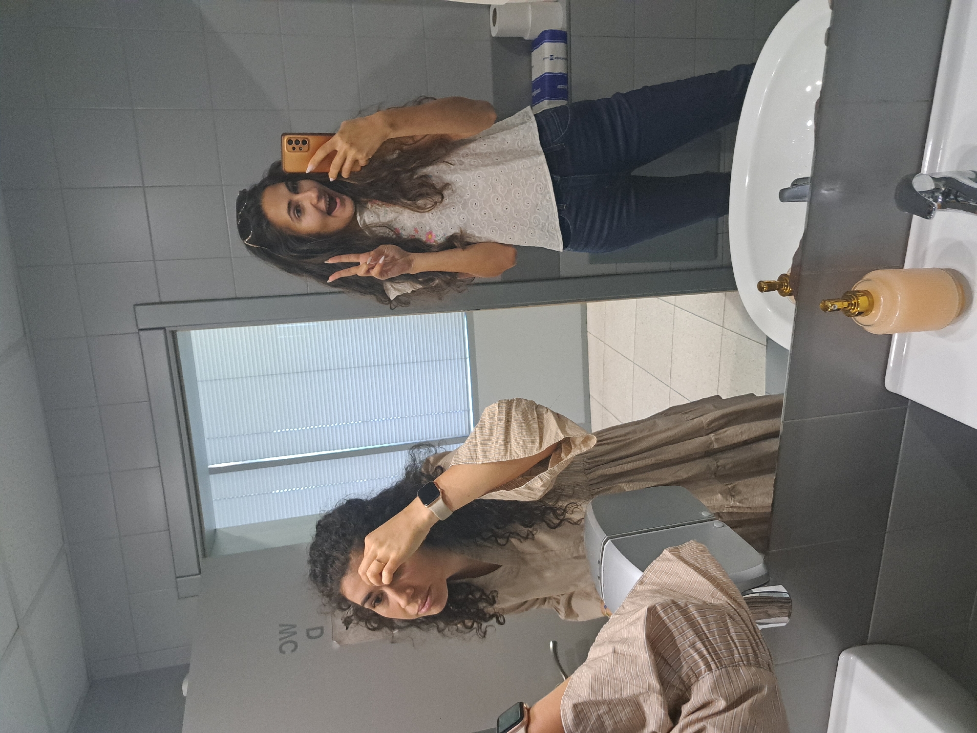 Two young women are in a public restroom. The woman on the left, with curly dark hair, is looking into the mirror and touching her face. She is wearing a beige dress with a tiered skirt and a striped shirt underneath. On her left wrist, she wears a smartwatch with a light-colored band. The woman on the right, also with long dark hair, is holding a smartphone and taking a selfie in the mirror. She is wearing a white top with a lace pattern and dark blue jeans. She is making a peace sign with her left hand and has her mouth open in a surprised expression. She is wearing sunglasses on top of her head. The restroom has gray tiled walls, a large mirror, a sink with a chrome faucet, a soap dispenser, and a hand dryer. In the background, there are toilet paper rolls and a box of tissues on a shelf. The floor is tiled with a light-colored pattern. The environment suggests a modern public facility, possibly in the city of Sofia, Bulgaria, given the context of the prompt.