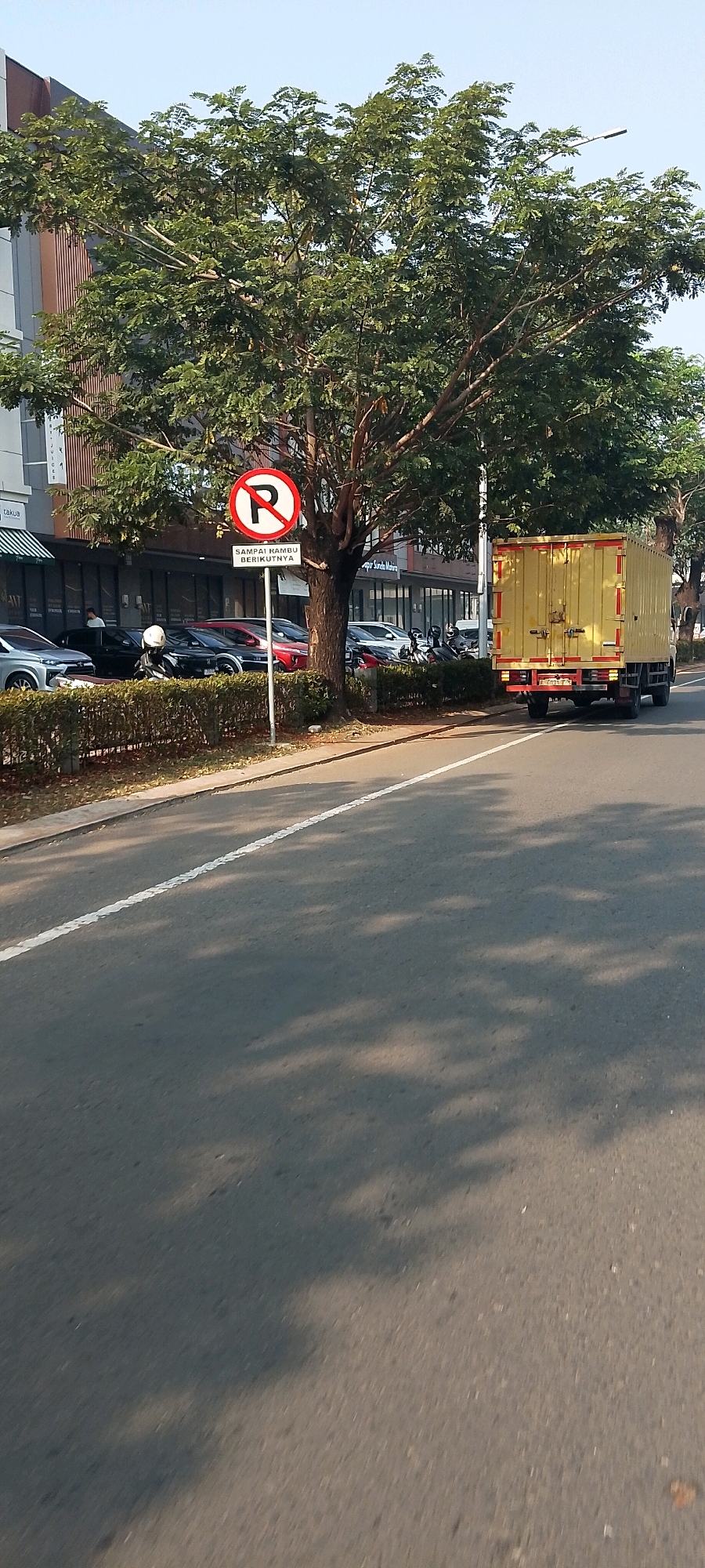 The image depicts a street scene in Gunung Putri, Indonesia, on a sunny day with clear skies. In the foreground, a gray asphalt road features a white lane marking and is dappled with shadows from overhead trees. Along the left side of the road, a low hedge separates the street from a sidewalk and a row of buildings. Parked cars and motorcycles are visible behind the hedge, along with a man standing near a building entrance.
A prominent circular red and white sign with a diagonal line through a 'P' symbol, indicating "No Parking," stands near the center of the image, accompanied by a smaller rectangular sign below it with Indonesian text. Behind the sign, a large tree with lush green foliage dominates the midground. Further to the right, a yellow box truck with red accents is driving away from the camera on the road. The buildings in the background appear to be commercial establishments with large glass windows and awnings. The overall environment suggests an urban or suburban commercial area.