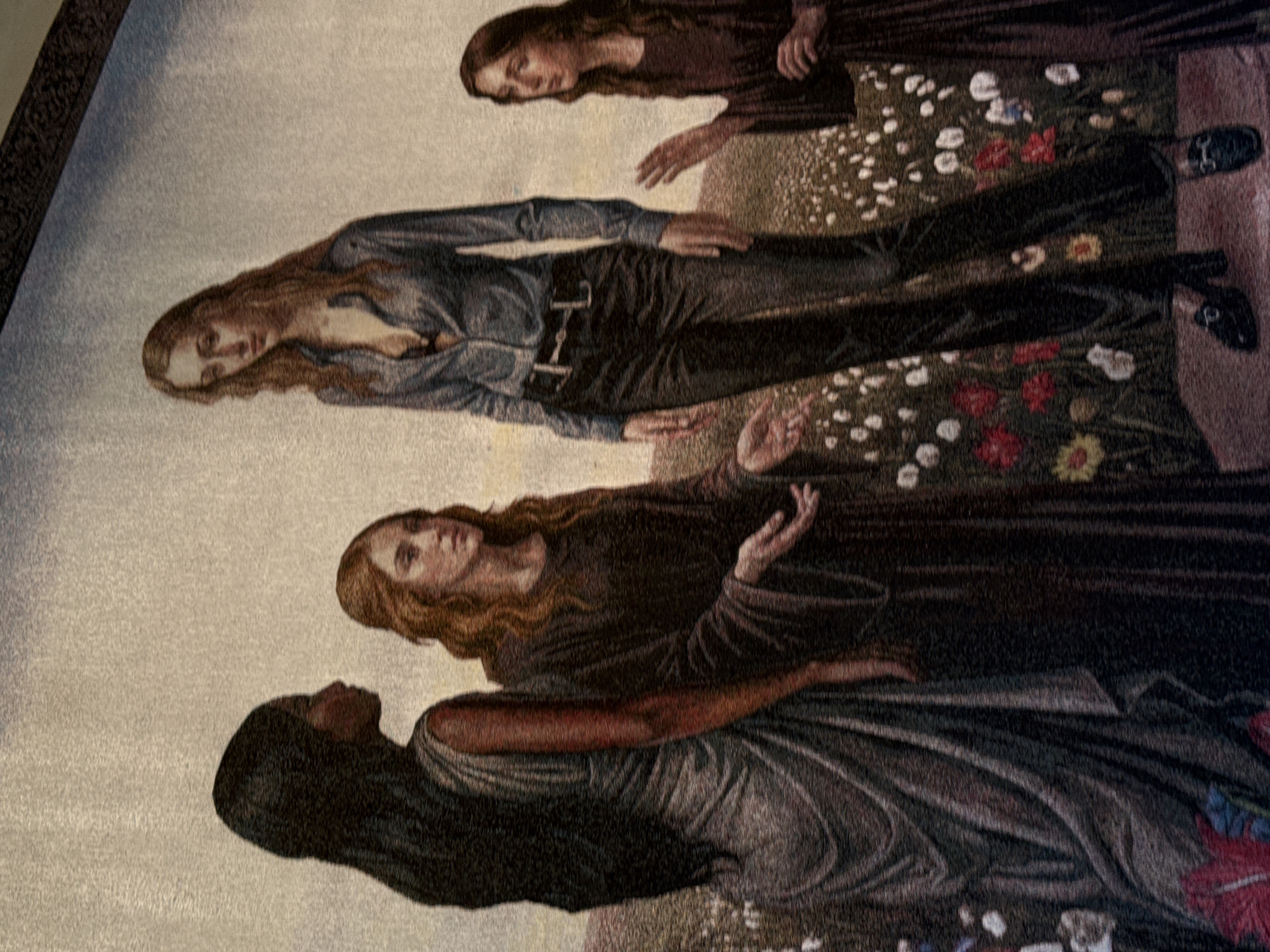 This appears to be a photograph of a tapestry depicting several women in a field.
From the bottom, a woman with dark hair is wearing a gown with dark shadows in it. Above her, a woman with shoulder-length brown hair wears a dark robe. A woman above her has longer brown hair and wears what looks like a denim top with black pants and a belt. At the top, a woman with long brown hair wears a dark dress.
The setting appears to be a grassy field with white flowers and some red flowers. It is unclear if this image is located in Milan, Italy.
