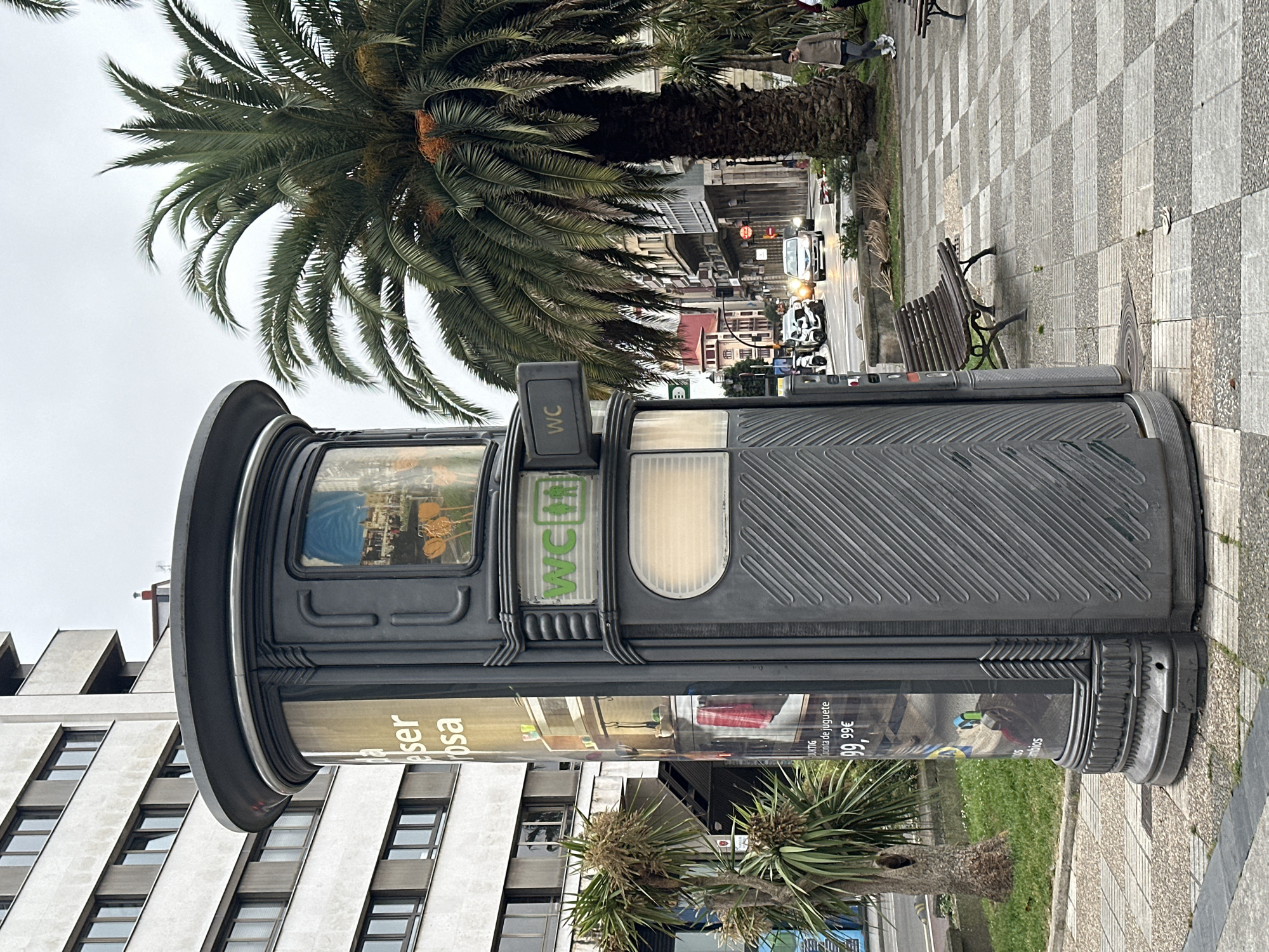 A public toilet (WC) stands on a checkerboard-paved sidewalk in Gijón/Xixón, Spain. It's cylindrical, dark gray with ridges, advertising displays, and a "WC" sign. A palm tree towers behind the structure. In the background, a street scene features buildings, cars, and pedestrians. A bench sits nearby on the sidewalk. The sky is overcast.