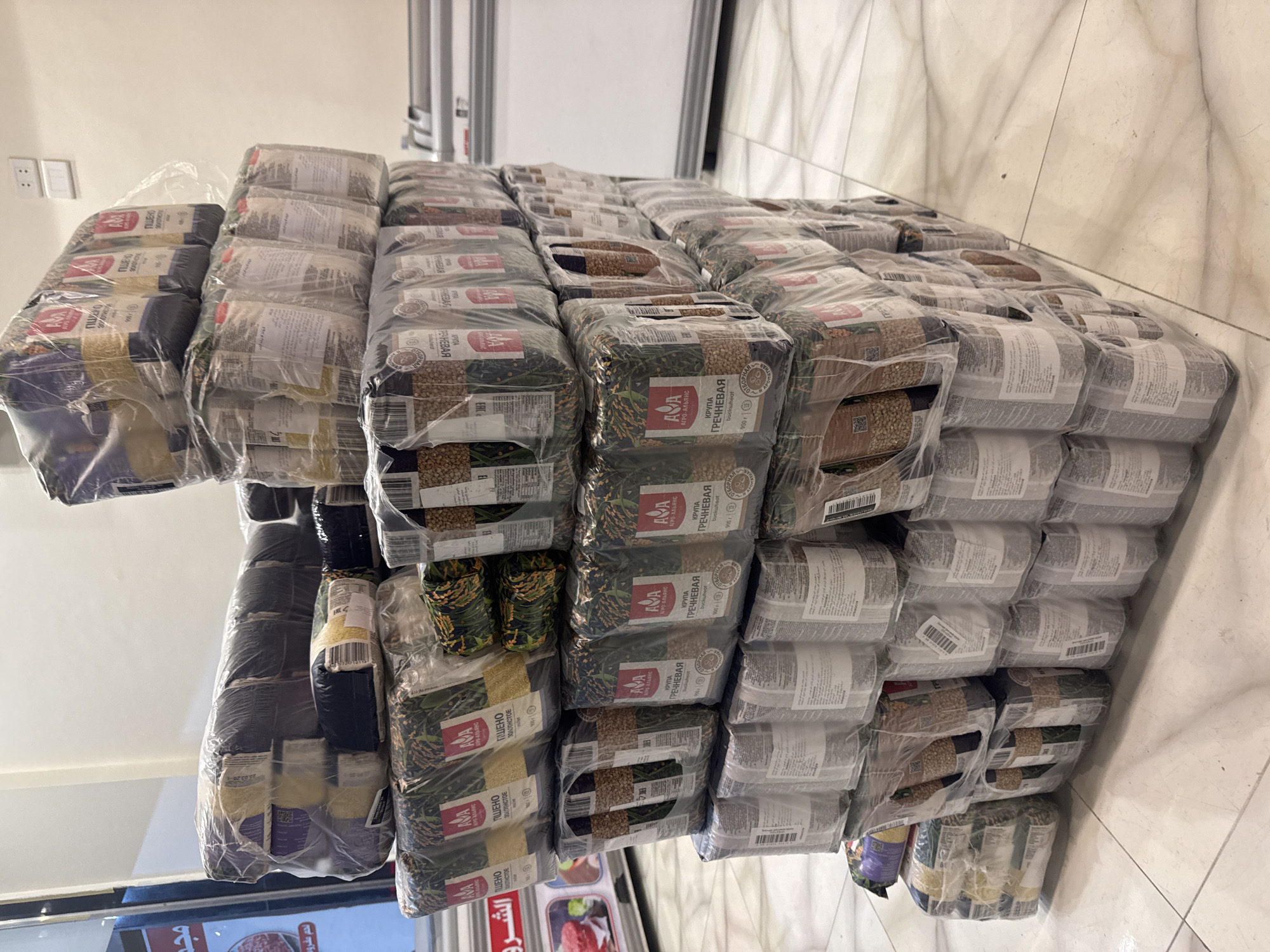 The image shows a large stack of packaged food items, likely grains, arranged on a tiled floor in an indoor location that could be a storage room, warehouse, or retail space in the city of Unknown, Unknown. The packages appear to be stacked in neat rows and columns. The labels on the packages have Russian text, suggesting that the product is intended for sale or distribution in a Russian-speaking market. The primary brands visible are "AVA" and "Agro-Alyans." Some of the packages contain buckwheat groats (ГРЕЧНЕВАЯ buckwheat), and some contain millet (ПШЕНО millet). The environment appears to be sterile and well-maintained, but there is no evidence of people or activity, and no interactions are taking place. The location is somewhat ambiguous.