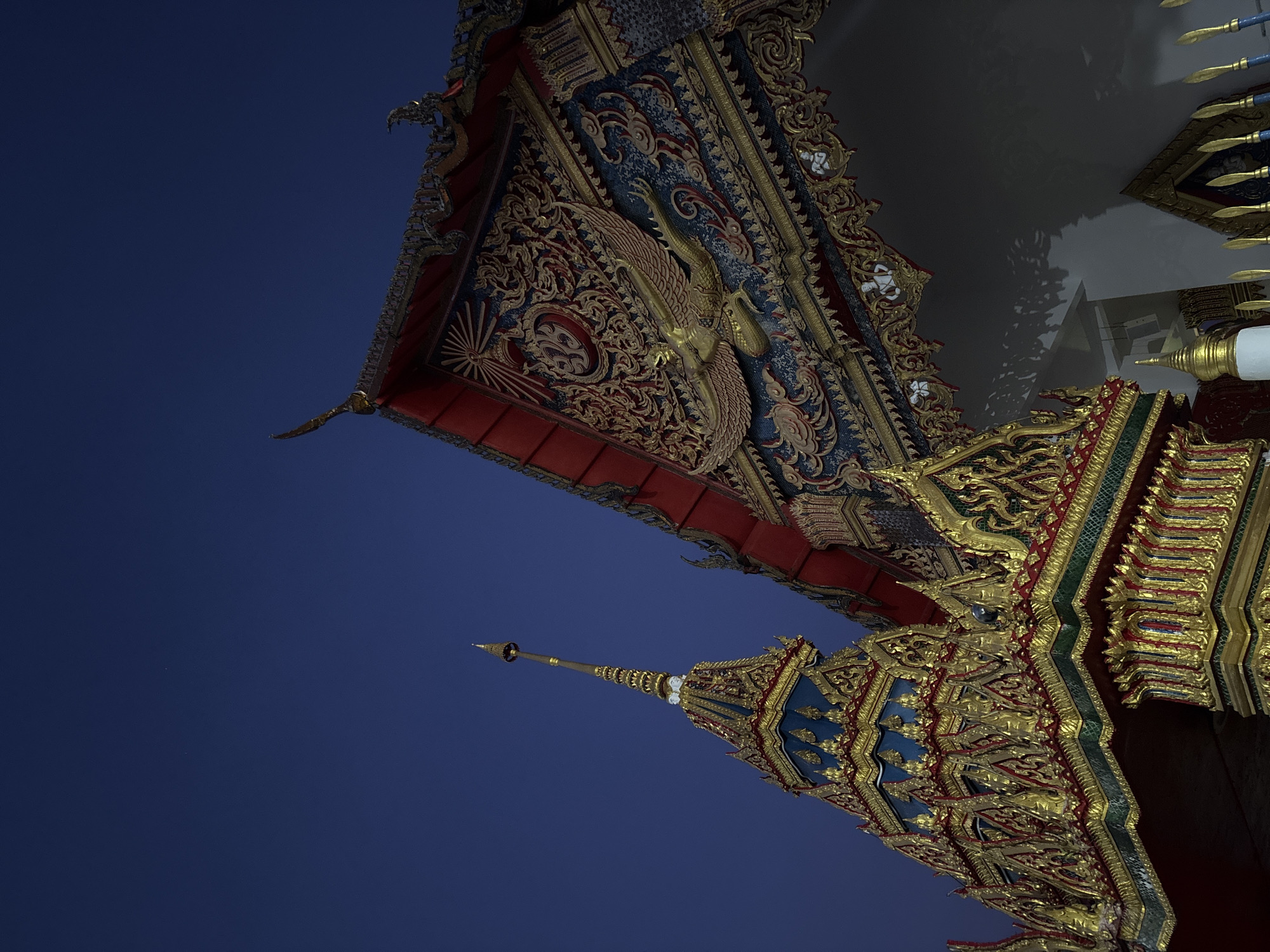 The image shows the intricate golden and ornate architecture of a Buddhist temple against a deep blue sky. The scene captures the details of the temple roof, adorned with traditional Thai designs and sculptures. The structure appears to be located in Phuket City Municipality, Thailand. There are no people visible in the image.