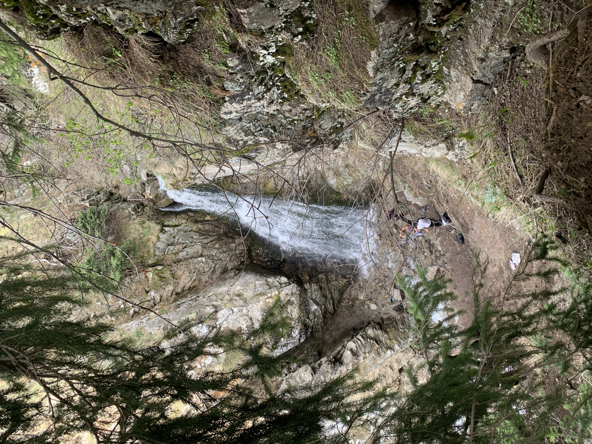 An aerial view of a waterfall, possibly near Almaty, Kazakhstan, is captured. The scene is framed by foliage, with a dense evergreen tree dominating the foreground. The waterfall cascades down a rocky terrain, surrounded by moss-covered rocks and sparse vegetation. On a patch of dry earth next to the waterfall, a group of people are present, with scattered clothing and bags. The overall environment suggests a natural, somewhat remote location.