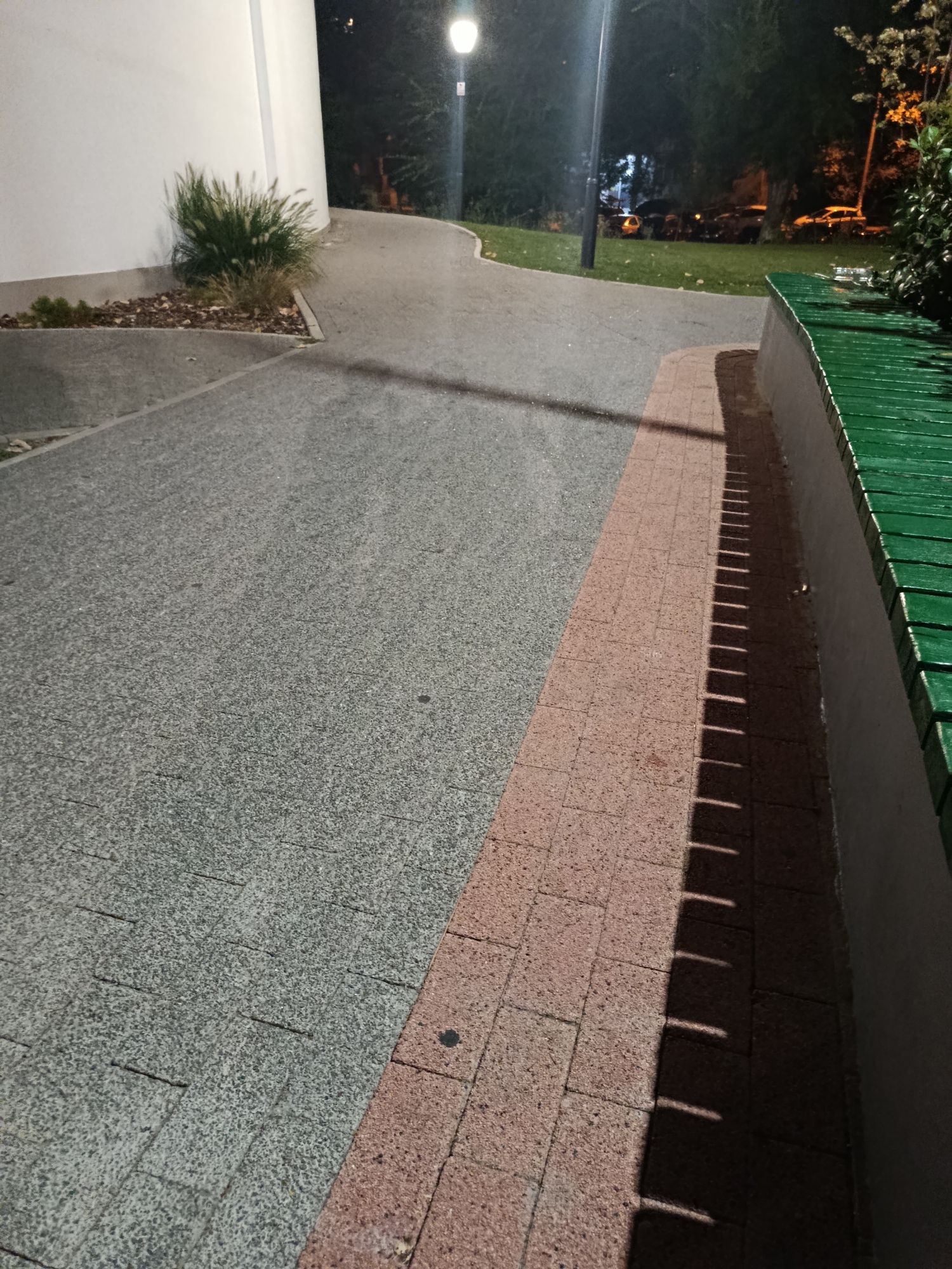 The image depicts an outdoor scene at night, likely in a park or public area in Chișinău, Moldova. The foreground is dominated by two pathways. The left pathway is paved with a coarse, grey, speckled material, possibly exposed aggregate concrete or granite chips, laid in rectangular slabs. Adjacent to this is a narrower pathway paved with reddish-brown rectangular bricks. The right side of the frame features a white wall, and above it, a portion of a green wooden bench is visible, its slats running horizontally. The bench casts a distinct shadow pattern onto the brick pathway, suggesting a light source from the right. In the background, a paved walkway curves away, illuminated by a streetlamp on a dark pole. Beyond the walkway, a grassy area is visible, with several cars parked on it or nearby under the cover of trees. The trees themselves are dark silhouettes against a faintly lit night sky, with some distant lights or possibly illuminated foliage suggesting an urban setting. A cluster of ornamental grass is planted near the white wall in the mid-left background. The overall impression is one of a quiet, possibly well-lit, public space at night.