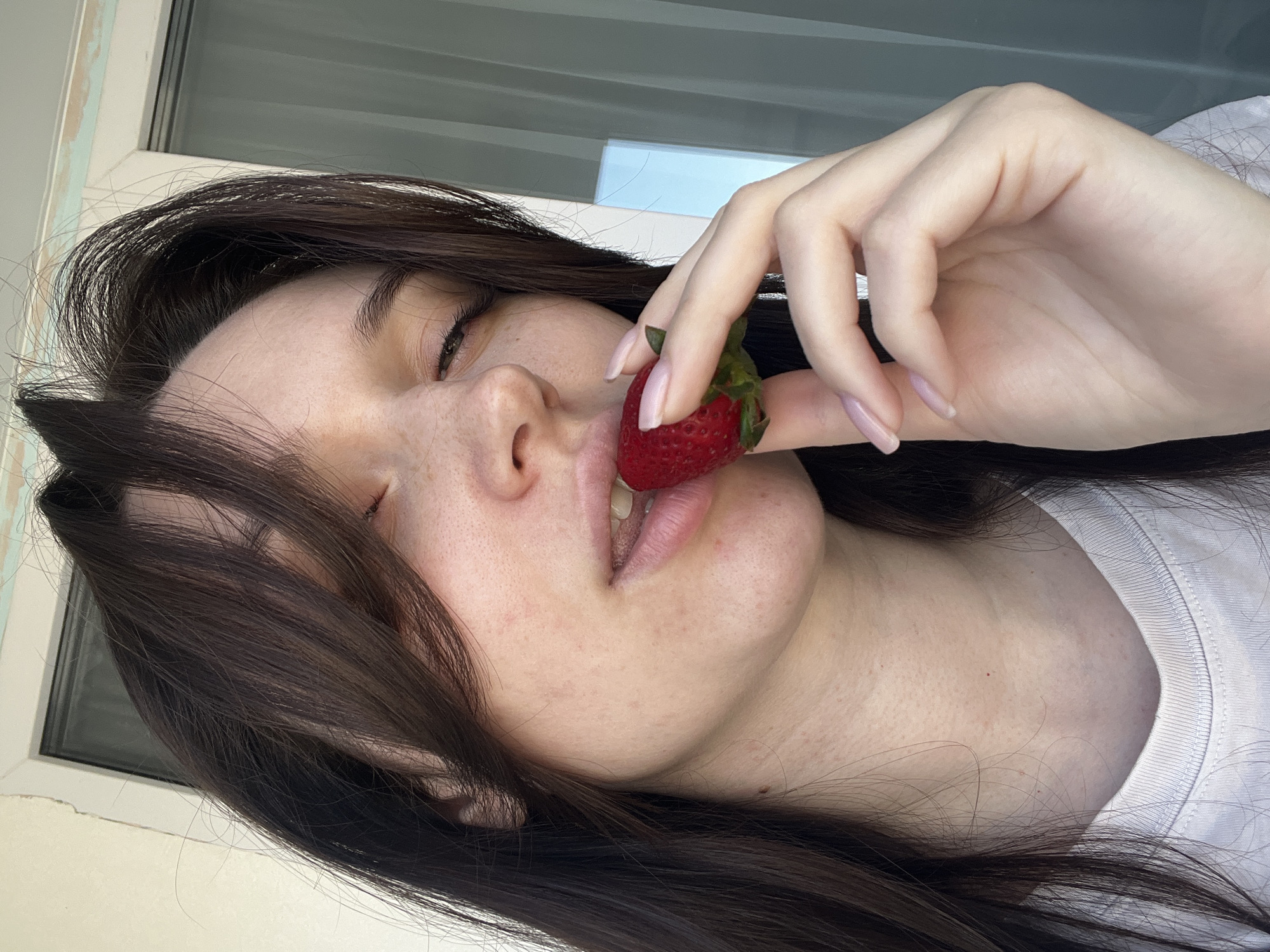 The image features a close-up portrait of a person, likely female, oriented in a way that suggests they are lying down or tilted, with their head resting against a surface. The person has long, dark brown hair that frames their face and extends across the bottom of the frame. Their eyes are closed, and they are holding a single, ripe red strawberry to their slightly parted lips with their right hand, which has light pink nail polish.
The setting appears to be indoors, near a window, providing natural light. Parts of a white or light-colored window frame are visible in the background, with a glimpse of what seems to be a grey building exterior and a sliver of blue sky through the glass. The person is wearing a white t-shirt, visible on the right side of the frame. Notable details on the skin include freckles and a few small moles. The overall atmosphere conveyed is one of calm, relaxation, or enjoyment, possibly related to the act of savoring the fruit. There is no visible text in the image. The location context provided for this image is Tyumen, Russia.