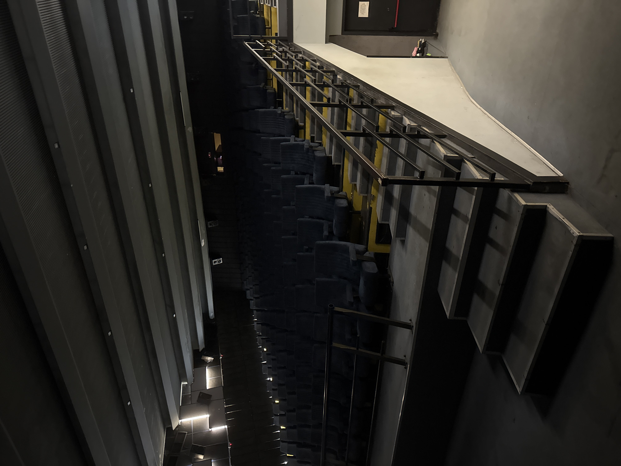 This is a low-angle, wide shot of an empty cinema auditorium. The focus is on the tiered seating and a wide staircase with a metal railing. The seats are upholstered in dark blue fabric and are arranged in rows that ascend from the lower level to the back of the auditorium. The staircase is painted black with yellow markings on the edges of the steps. The walls of the auditorium are dark gray, and the ceiling features parallel, dark gray beams with integrated lighting fixtures. In the background, there are illuminated panels on the wall and speakers mounted near the ceiling. A green door with a sign is visible on the right side of the image. The lighting is dim, creating shadows that emphasize the architectural features of the space. The overall impression is one of a quiet, unoccupied cinema. The image does not provide any specific information about the city of Blagoevgrad, Bulgaria.