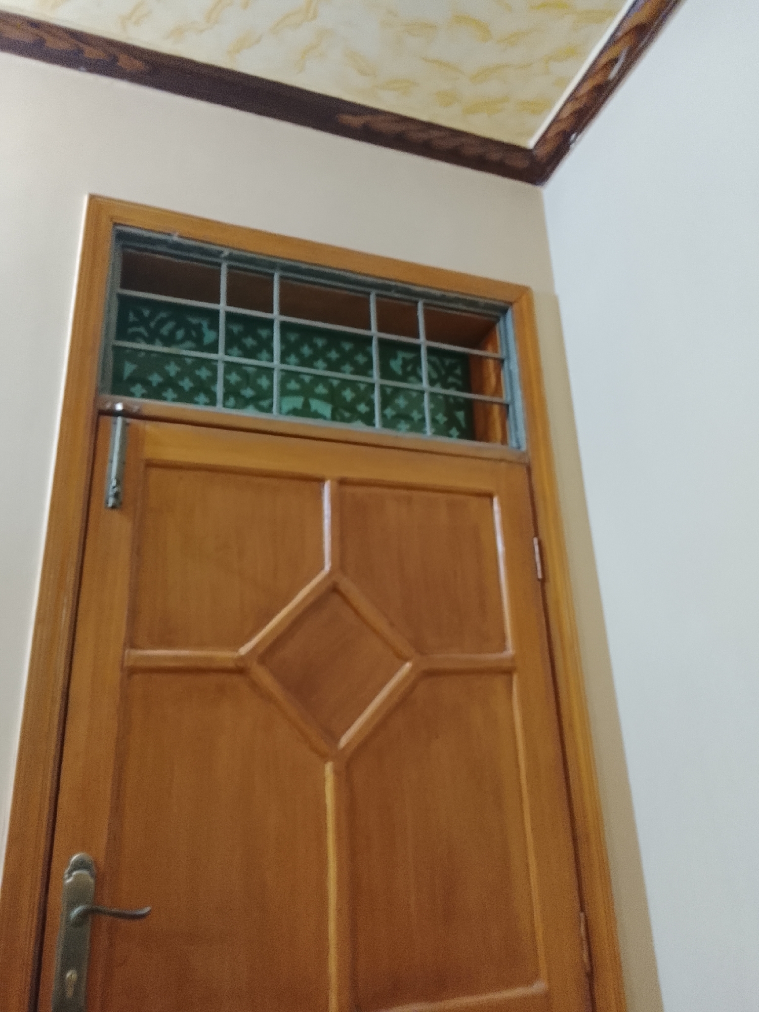 A medium-shot, upward-angled photograph features a wooden door with a metal-grilled transom window, likely situated within a building in Multan, Pakistan. The door is primarily golden brown, with a decorative diamond shape at its center. Above the main door panel, a rectangular transom window with a grid-like metal frame contains a translucent green patterned screen. The door has a silver-colored lever handle on the lower left. The door is framed by a wooden architrave of similar color to the door, slightly lighter than the door itself. The surrounding walls are painted in a pale cream or off-white color. The ceiling above the door has a decorative border of dark brown, with a lighter brown and yellow patterned design on the ceiling itself. No people are visible.
