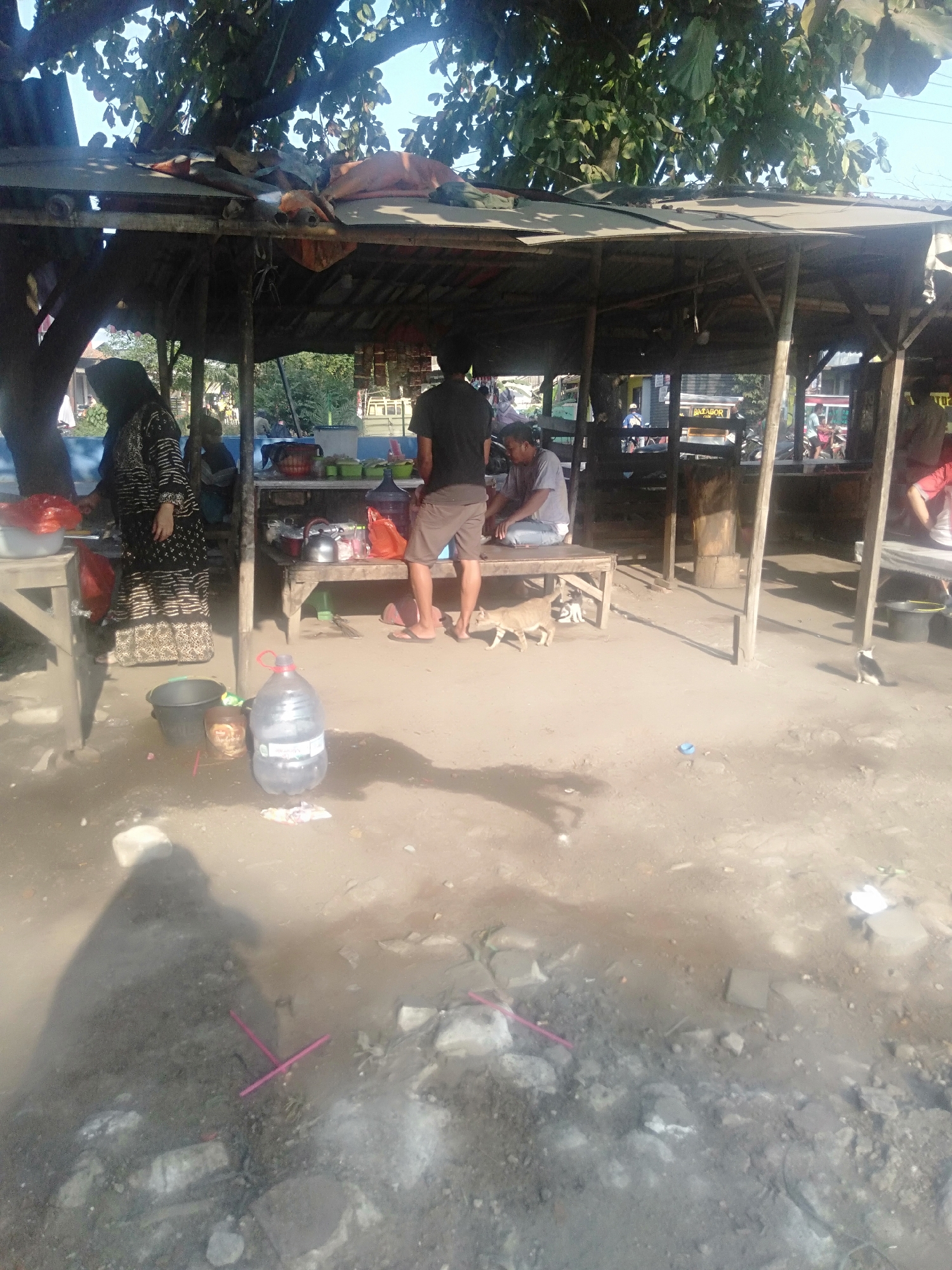 The image shows an outdoor scene with a rustic, covered structure that appears to be a food stall or a small shop. The structure is supported by wooden posts and has a corrugated roof, with some makeshift additions. Several people are present. On the left, a person wearing a headscarf and a patterned dress stands near a table, with red plastic bags on it. In the center, a person in a black t-shirt and shorts stands, looking down, with a table in front of them holding various items, including a kettle, bottles, and plastic containers. To the right of this central figure, another person is seated on a low bench. A dog and a small cat are visible on the sandy ground in front of the stall. Various objects are scattered around the area, including buckets, large plastic water bottles, and debris. The ground is uneven and sandy. In the background, there are trees and glimpses of other structures and activity, suggesting a rural or semi-urban setting. The lighting indicates it is daytime, with sunlight casting shadows. The scene is set in Sukadami, Indonesia.