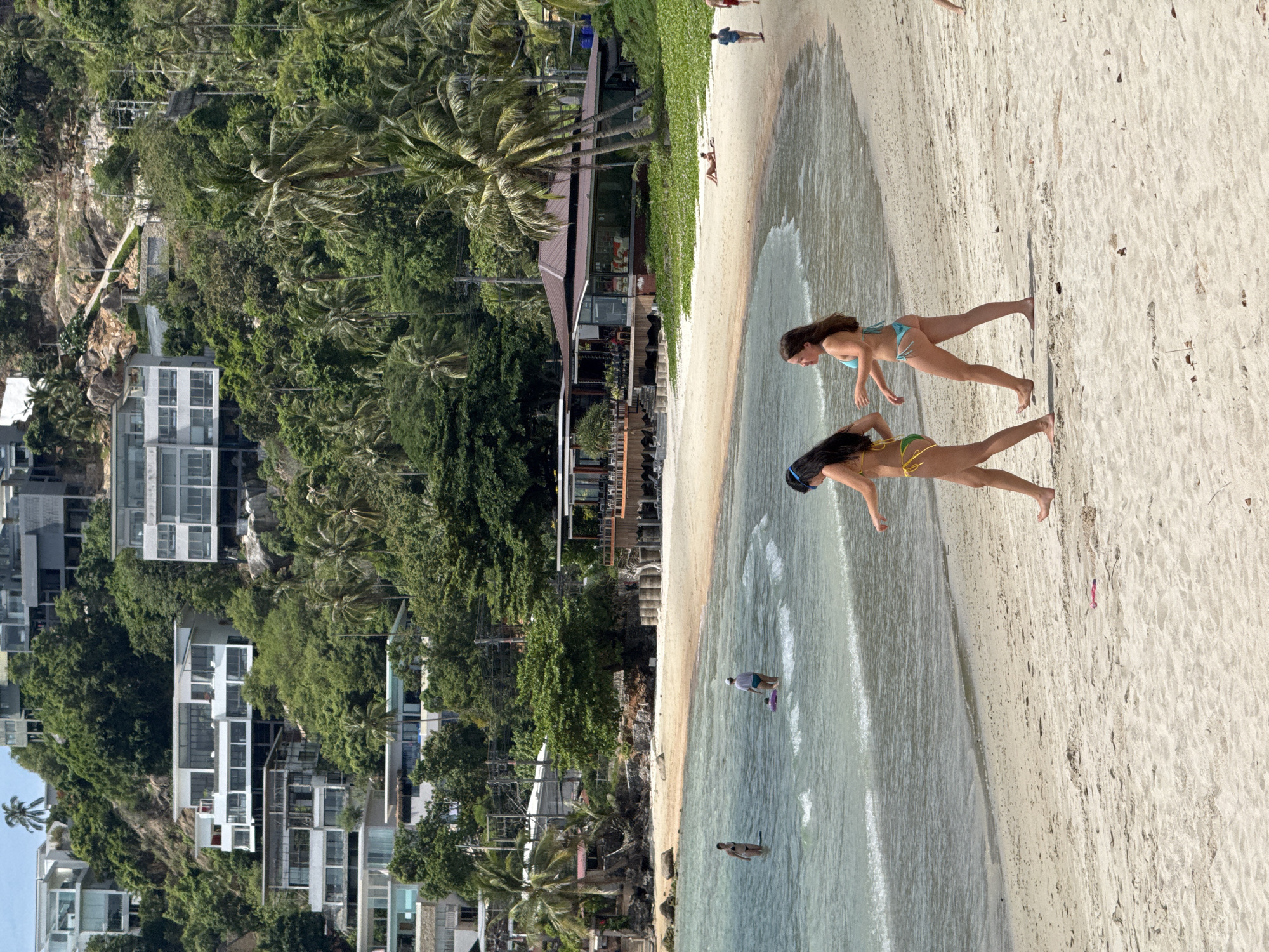 This image depicts a tropical beach scene on the island of Ko Samui, Thailand, likely within the Ko Samui District. In the foreground, two young women in bikinis are walking on the sandy beach. The woman on the left wears a yellow bikini with green trim, while the woman on the right wears a blue bikini. The water is calm and shallow, with gentle waves lapping at the shore.
In the mid-ground, several people are visible in the water. One person, wearing a white cap, stands in the water with a purple flotation device. Another person, wearing a dark bikini, stands further out in the water. A small, bright pink object, possibly a child's toy, is submerged near the person in the white cap.
Behind the beach, there is a long, wooden building with a brown roof, likely a restaurant or bar, with outdoor seating visible. Palm trees and lush green vegetation cover a steep hillside behind the building.
In the background, modern white buildings with multiple levels and large windows are constructed on the hillside, indicative of resort accommodations or private residences. The architecture suggests a contemporary design that blends with the natural environment. Power lines are visible running along the hillside, connecting the structures. The sky is a clear, bright blue. The overall impression is a sunny day at a developed tourist destination.