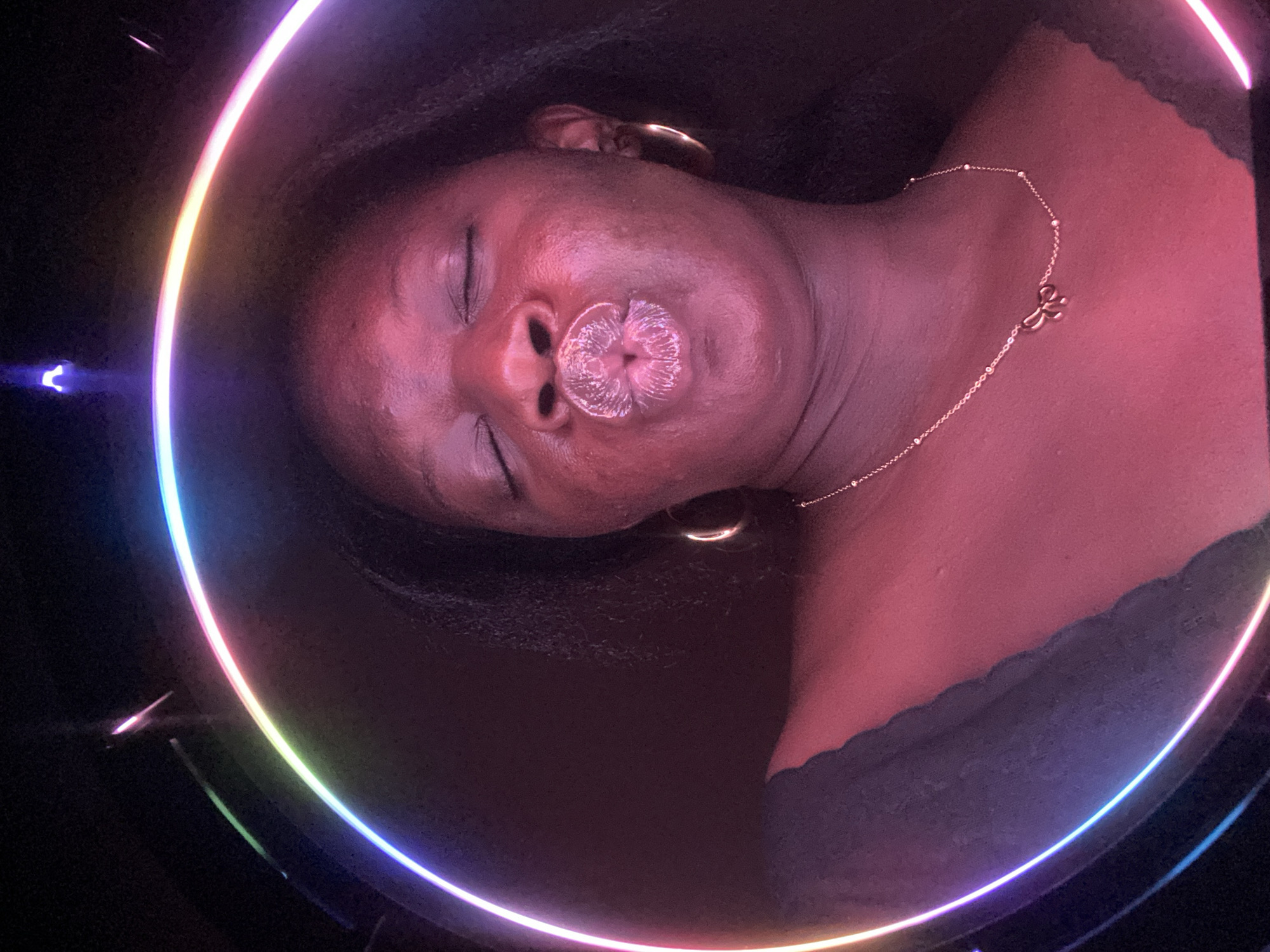 A medium shot captures a woman with dark skin and closed eyes, illuminated by a circular neon light. Her lips are prominent and appear decorated. She wears hoop earrings and a delicate necklace with a bow-shaped pendant. The background is dark and indistinct. There is no indication of a specific location in the city of Maigizo, Nigeria.