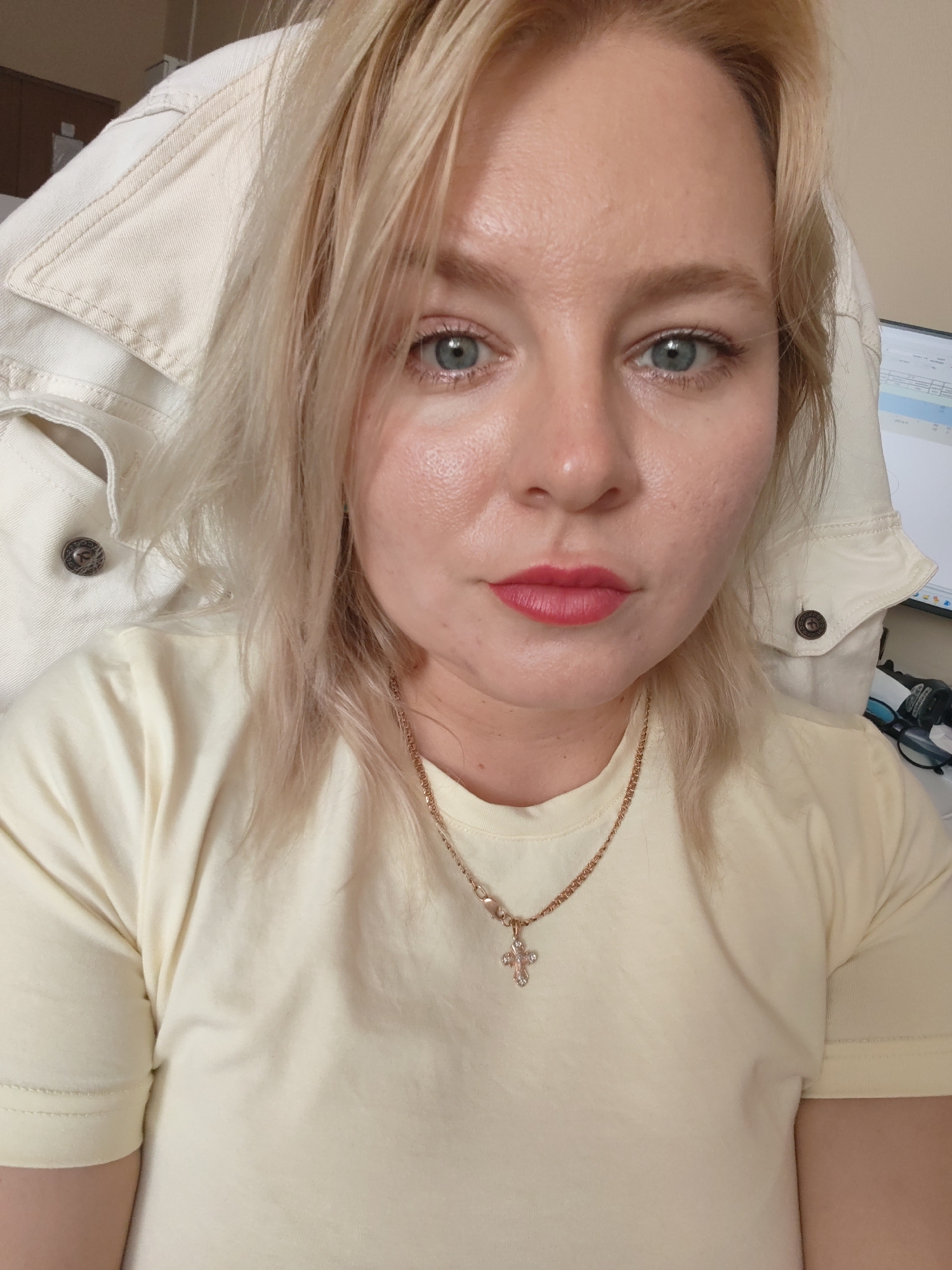 A woman with blonde hair, blue eyes, and red lipstick is looking at the camera. She is wearing a light yellow t-shirt and a cream-colored jacket. She is also wearing a gold chain with a cross pendant. The background is blurry, but it appears to be an office setting. There is a computer monitor and a pair of glasses visible in the background. The image was taken in Izhevsk, Russia.