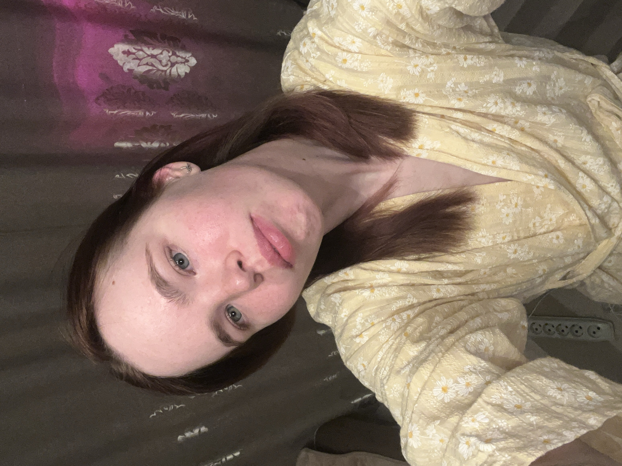 A medium shot depicts a young woman with fair skin, light blue eyes, and auburn hair. She is positioned at an oblique angle to the camera, lying down. Her facial expression is neutral. She is wearing a light yellow dress with a white daisy floral pattern. The background consists of bedsheets, one section displaying a pink and white floral pattern, and another appearing grey. In the lower right corner of the image, a white electrical outlet strip is partially visible. There is no verifiable evidence in the image to confirm the location in Chaikovsky, Russia.