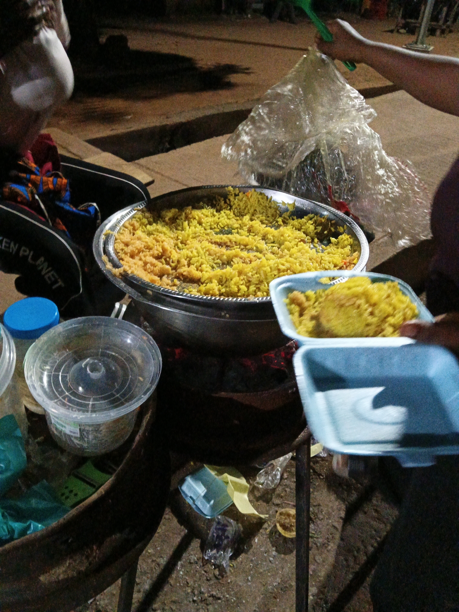 An outdoor scene in Bukuru, Nigeria, at night shows a person serving a large metal bowl of yellow rice. The rice is being scooped into a blue styrofoam container held by another person. A plastic bag, likely for packaging, is being manipulated with a green utensil. In the foreground, a blue plastic container lid and a clear plastic container are visible. The ground is dirt and debris.