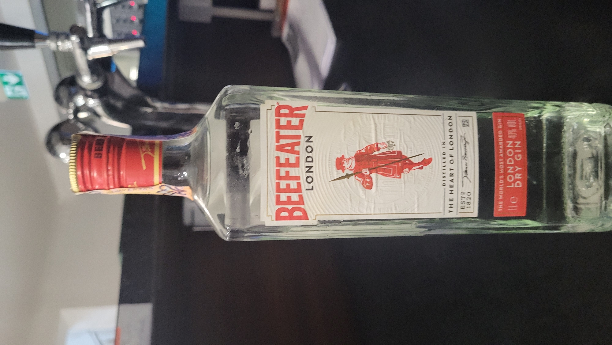 The image shows a full, unopened bottle of Beefeater London Dry Gin. The bottle is clear glass and rectangular in shape, with a red and gold foil cap and a white label. The label features the Beefeater logo, the words "LONDON" and "DISTILLED IN THE HEART OF LONDON," a depiction of a Yeoman Warder, and "ESTD 1820." Below this is a red band with "THE WORLD'S MOST AWARDED GIN," "1le LONDON 40% VOL," and "DRY GIN." The bottle is standing upright on a dark surface, possibly a bar or counter. In the background, out of focus, are what appear to be beer taps and possibly some documents on a desk. The lighting is artificial. The location is identified as a bar in Prešov, Slovakia. There are no people visible in the image.