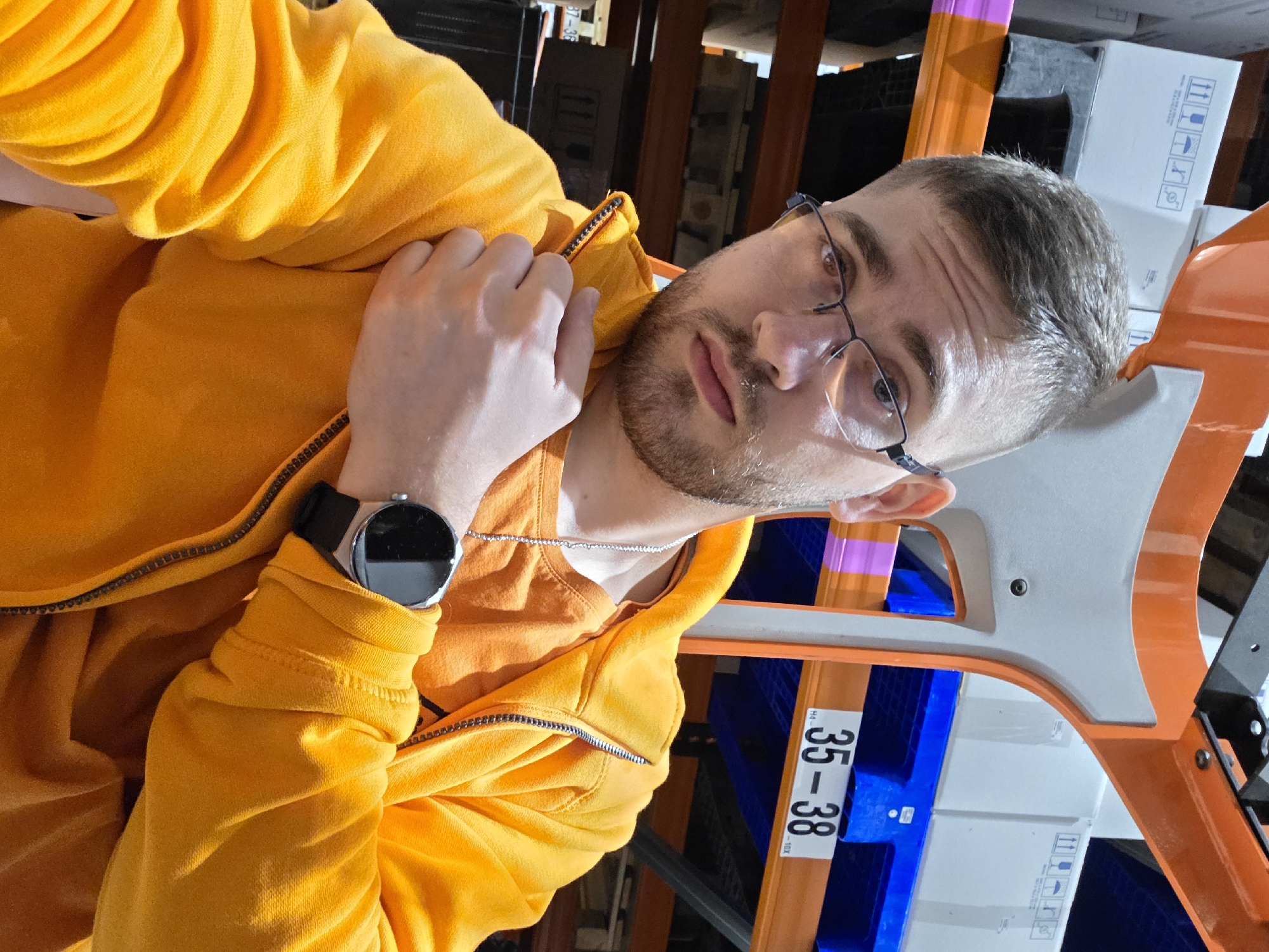 A man wearing glasses, a yellow jacket, and a wristwatch is in an industrial location with metal racking visible. The racking includes blue shelving with visible boxes, and orange support beams marked with "35-38". The location is not identifiable as being in Plovdiv, Bulgaria based on the available visual information.