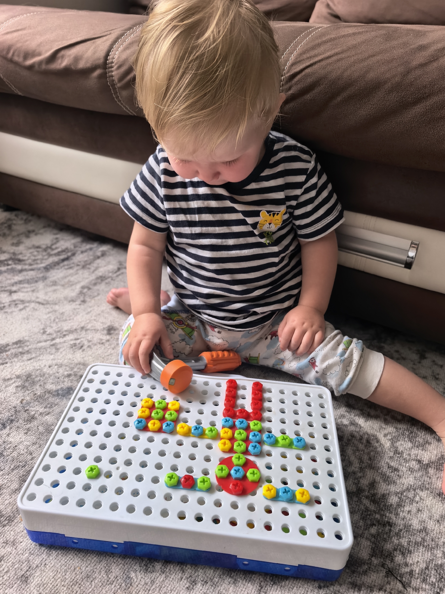 In a residential setting, a young blonde-haired child with fair skin is sitting on a patterned carpet and engaging with a toy construction set. The child is wearing a black and white striped t-shirt with a tiger graphic and light-colored pants adorned with a cloud and animal print. The child's right hand is holding an orange and gray toy tool, and their gaze is directed downwards at the toy. The construction set consists of a white pegboard with numerous holes, a blue base, and various colorful plastic pegs in shades of red, yellow, green, and blue, which have been partially arranged on the board to form abstract shapes. A brown sofa is visible in the background. The image does not provide any information that would allow for the identification of its location in Sterlitamak, Russia.