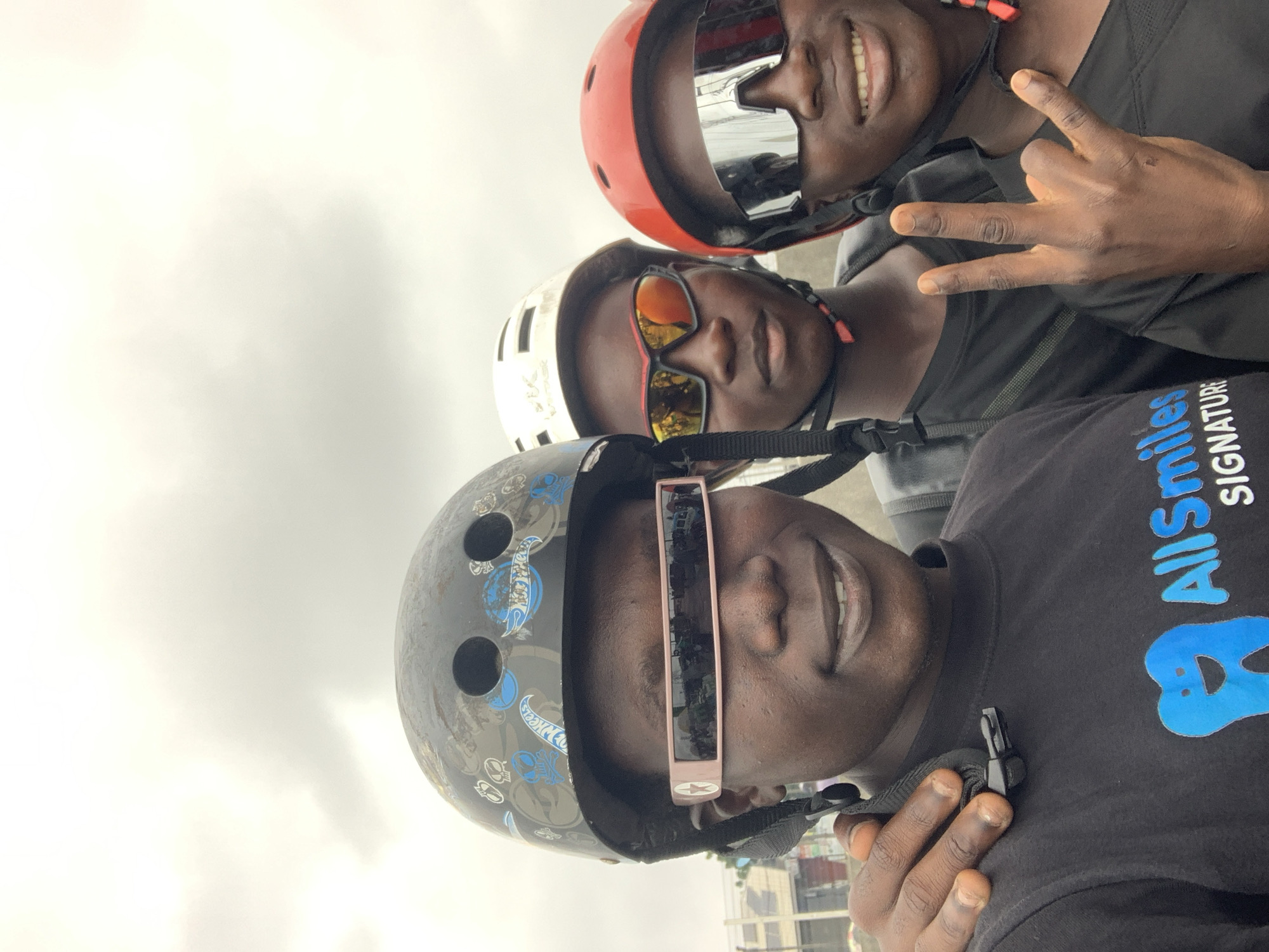 Three young individuals, appearing to be in their late teens or early twenties, are captured in a close-up, waist-up photograph outdoors. They are all wearing helmets and sunglasses, suggesting an activity that requires protective gear, possibly cycling or skateboarding. The person on the left is wearing a black helmet with a patterned design, and a black t-shirt with a blue logo featuring a stylized tooth and the text "AllSmiles SIGNATURE". They are also wearing distinctive pink-framed futuristic-looking sunglasses. The individual in the middle wears a white helmet and dark sunglasses with orange-tinted lenses. The person on the right is wearing a red helmet and large, silver-mirrored sunglasses. Their right hand is raised, forming a peace sign gesture with their index and middle fingers extended, while their thumb and pinky fingers are also extended. The background is slightly blurred, showing hints of urban infrastructure, possibly a street or market area, with a cloudy sky overhead. The overall lighting is soft and diffused, indicating an overcast day. The individuals are positioned close together, with the person on the left's arm resting on the shoulder of the person in the middle, indicating camaraderie. The scene appears to be set in a vibrant, outdoor urban environment, consistent with the description of Lekki, Nigeria, known for its lively atmosphere and recreational spaces.