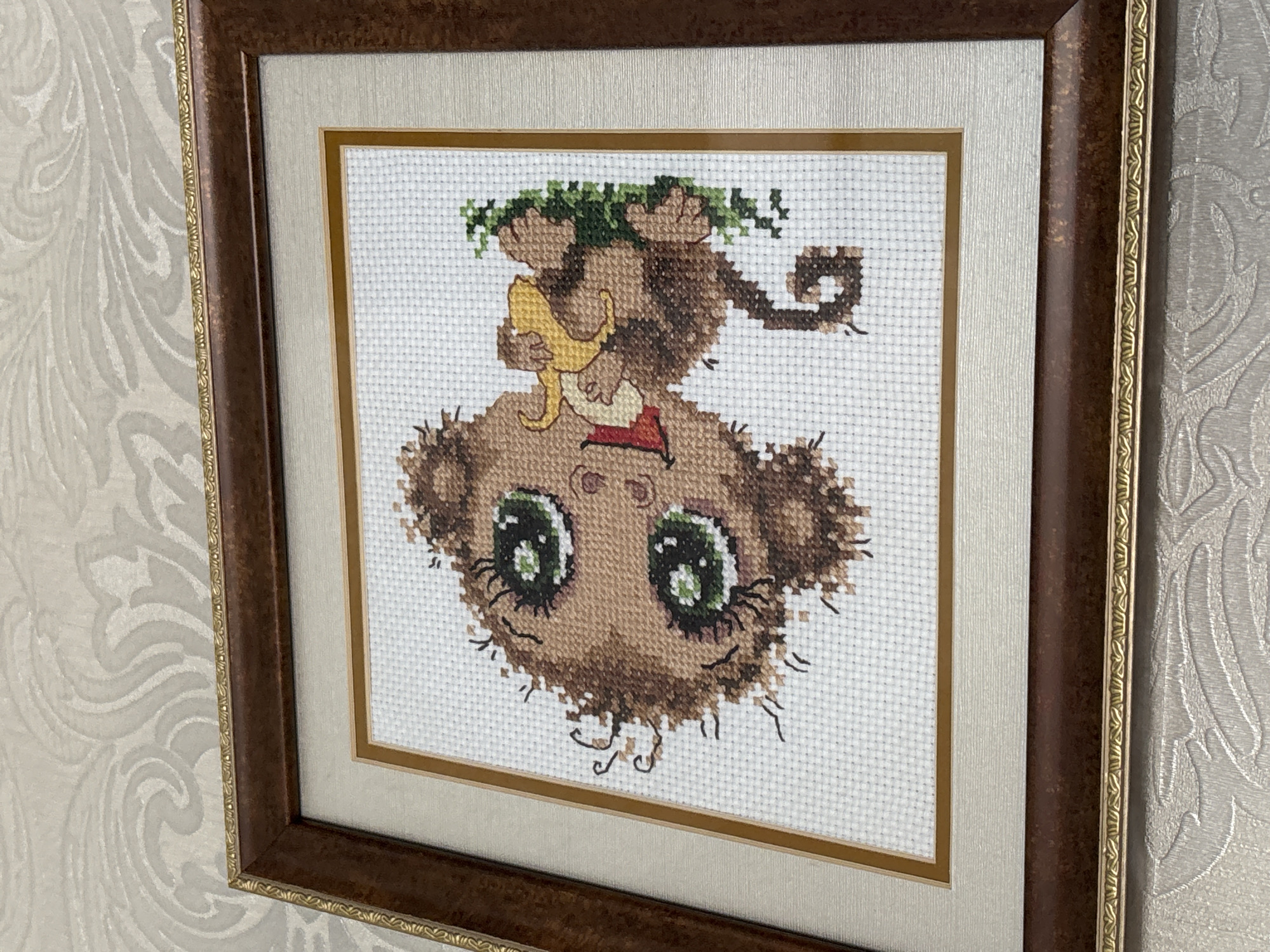 The image displays a framed cross-stitch artwork of a cartoon monkey holding a banana. The monkey is depicted with large green eyes, brown fur, and a smiling expression. It is sitting on a patch of green grass. The artwork is matted and framed in a dark brown wooden frame with gold trim. The frame is hung on a wall with textured wallpaper. The scene is an indoor setting, likely a home. The image does not contain any information about its location in Pavlodar, Kazakhstan.