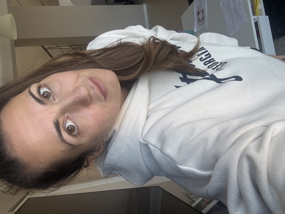 The image depicts a close-up, selfie-style photograph of a single female subject within a modern residential interior in Varna, Bulgaria. The woman, appearing to be in her late 20s to early 40s, has long, light brown hair, some of which is pulled back while other strands fall over her left shoulder. Her wide, brown eyes are directed at the camera, and she displays a neutral to slightly smiling expression with faintly parted lips. Her complexion is fair, with subtle freckling visible on her forehead and nose. She wears a light cream or off-white hooded sweatshirt featuring partial dark lettering on the chest that reads "STUSSY GEORGIA." A small, dark stud earring is visible in her left ear.
The background, partially blurred, reveals a well-lit indoor environment with natural light entering from the left. Visible elements include a light-colored ceiling pendant lamp in the upper right quadrant. A modern staircase with thin metal railings ascends in the background, suggesting a multi-level dwelling. To the lower right, a white table or desk surface is visible, holding papers and what appears to be a small decorative item with red accents. A dark, reflective surface is partially visible on the far left of the frame. The woman is actively holding the camera or phone to capture this self-portrait.