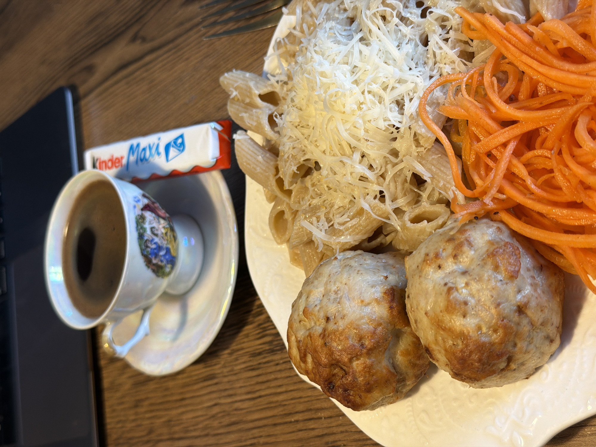 The image displays a meal and beverages arranged on a wooden table. In the foreground, a white, rectangular plate holds two large, lightly browned, spherical patties, likely meat or vegetable based. Adjacent to the patties, a portion of penne pasta, coated in a light sauce, is generously topped with grated white cheese. A separate pile of bright orange, julienned carrots is situated on the plate, next to one of the patties and the pasta. A silver fork is partially visible on the right side of the plate.
In the background, to the left, a small white iridescent teacup, adorned with a multi-colored classical scene, containing dark coffee, rests on a matching saucer. To the right of the teacup, a white and red "Kinder Maxi" chocolate bar in its packaging is visible. The top-left corner of a dark-colored laptop computer is partially seen, positioned behind the teacup. The entire arrangement is situated on a wooden surface exhibiting a distinct grain pattern, suggesting an indoor setting.
There is no visual information within the image to identify its specific location as Perm, Russia, or any other city or country.