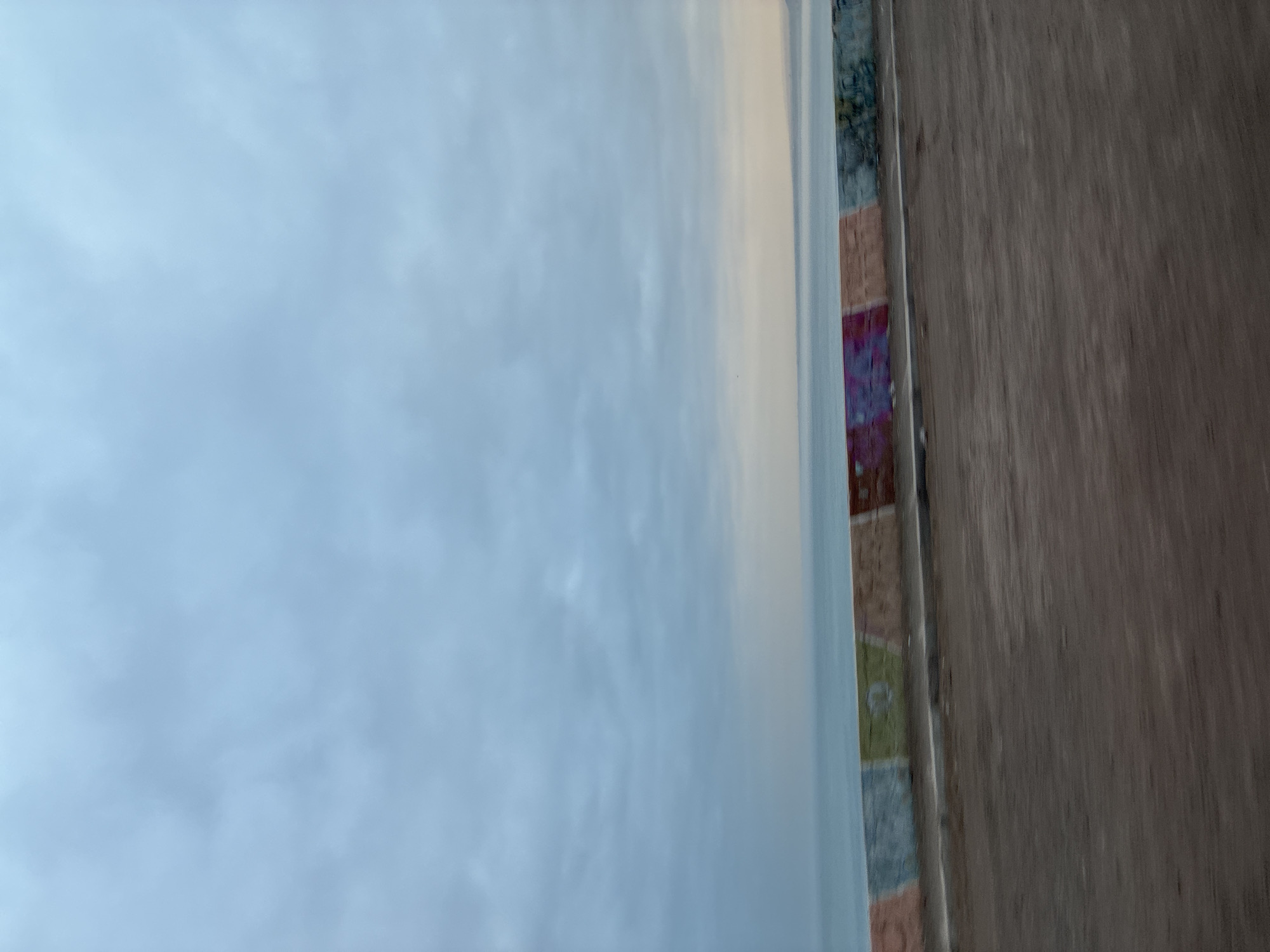 The image displays an outdoor scene under an overcast or partly cloudy sky. The upper portion of the frame is dominated by the sky, characterized by soft, pale blue hues and diffuse white or light grey cloud formations, suggesting a daytime setting with indirect sunlight.
Stretching horizontally across the lower part of the sky, there is a distinct band of decorative mosaic tiles. These tiles are small and vary in shape and color, featuring a muted palette of blues, greens, terracotta, browns, and subtle hints of purple, forming an irregular, textured pattern. This mosaic appears to be an architectural detail, possibly a border on a building or a decorative element at the edge of a structure.
To the right of the mosaic band, and occupying the lower right section of the image, a textured wooden surface is visible. This surface exhibits clear vertical wood grain and appears weathered, indicative of an exterior wooden wall or panel. The perspective suggests a view looking out from or past a building element, with the wooden surface being closer to the viewer than the mosaic and the sky.
No people or animals are present, and no specific activity is depicted. The scene offers a quiet observation of architectural textures against a subdued sky, characteristic of a coastal or urban setting such as Arica, Chile. There is no visible text within the image.